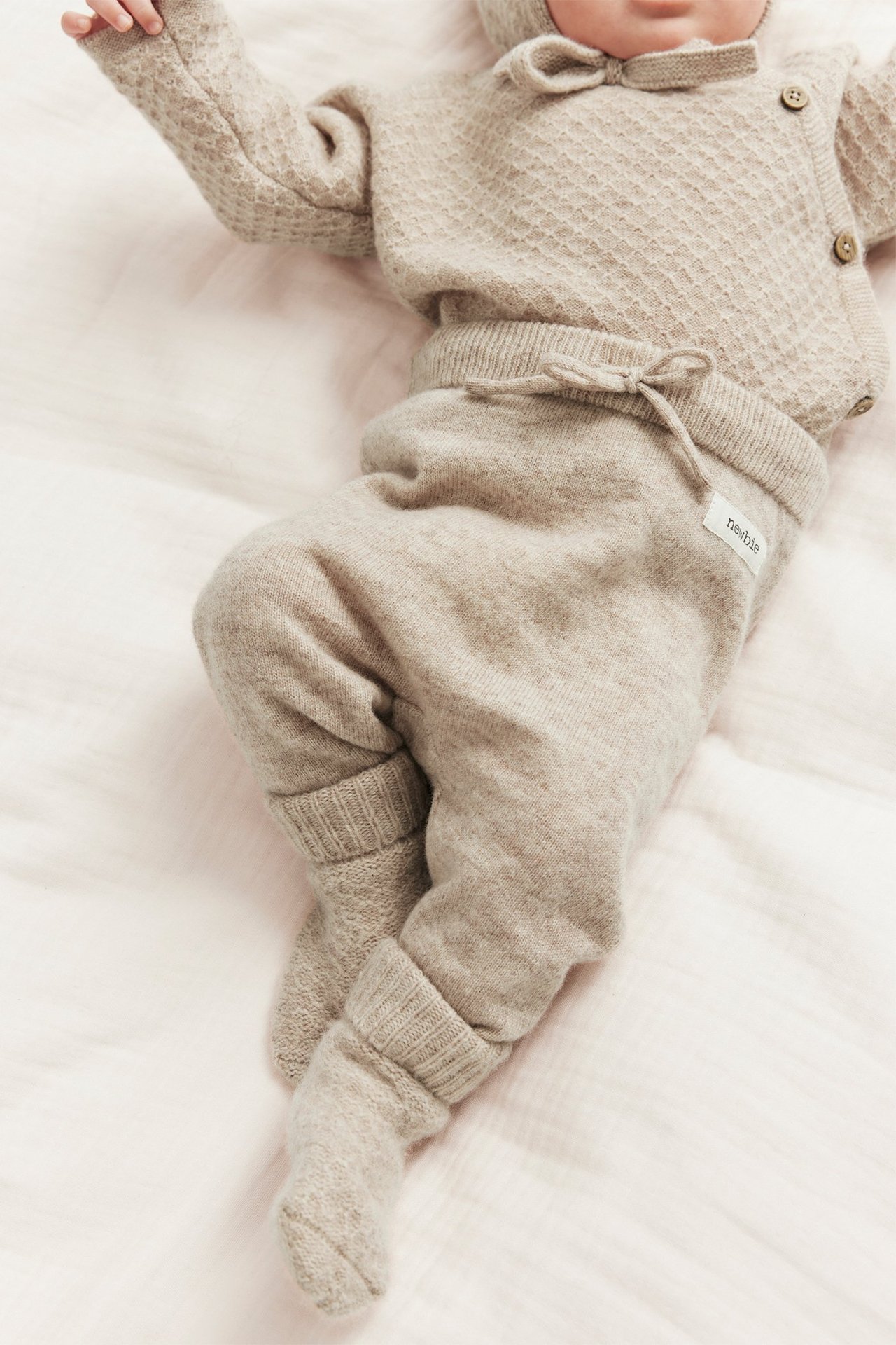 Baby wears beige melange wool-cashmere knit pants with drawstring, matching cardigan and bonnet. Top-down view.
