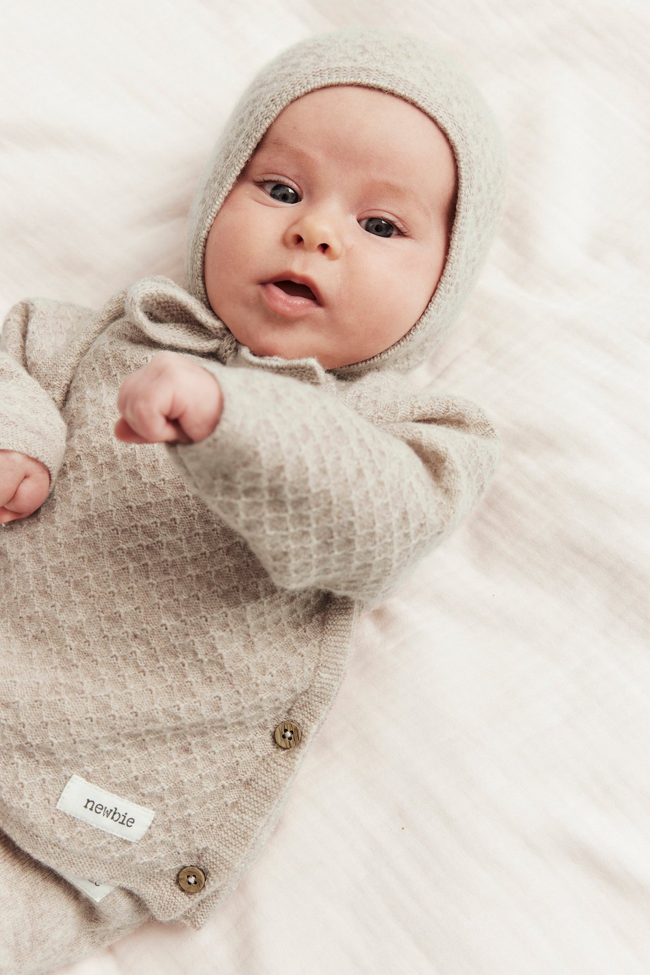Nahaufnahme eines Babys in beigemelange Strick-Woll-Kaschmir-Outfit mit Seitenknöpfen und passender Mütze.