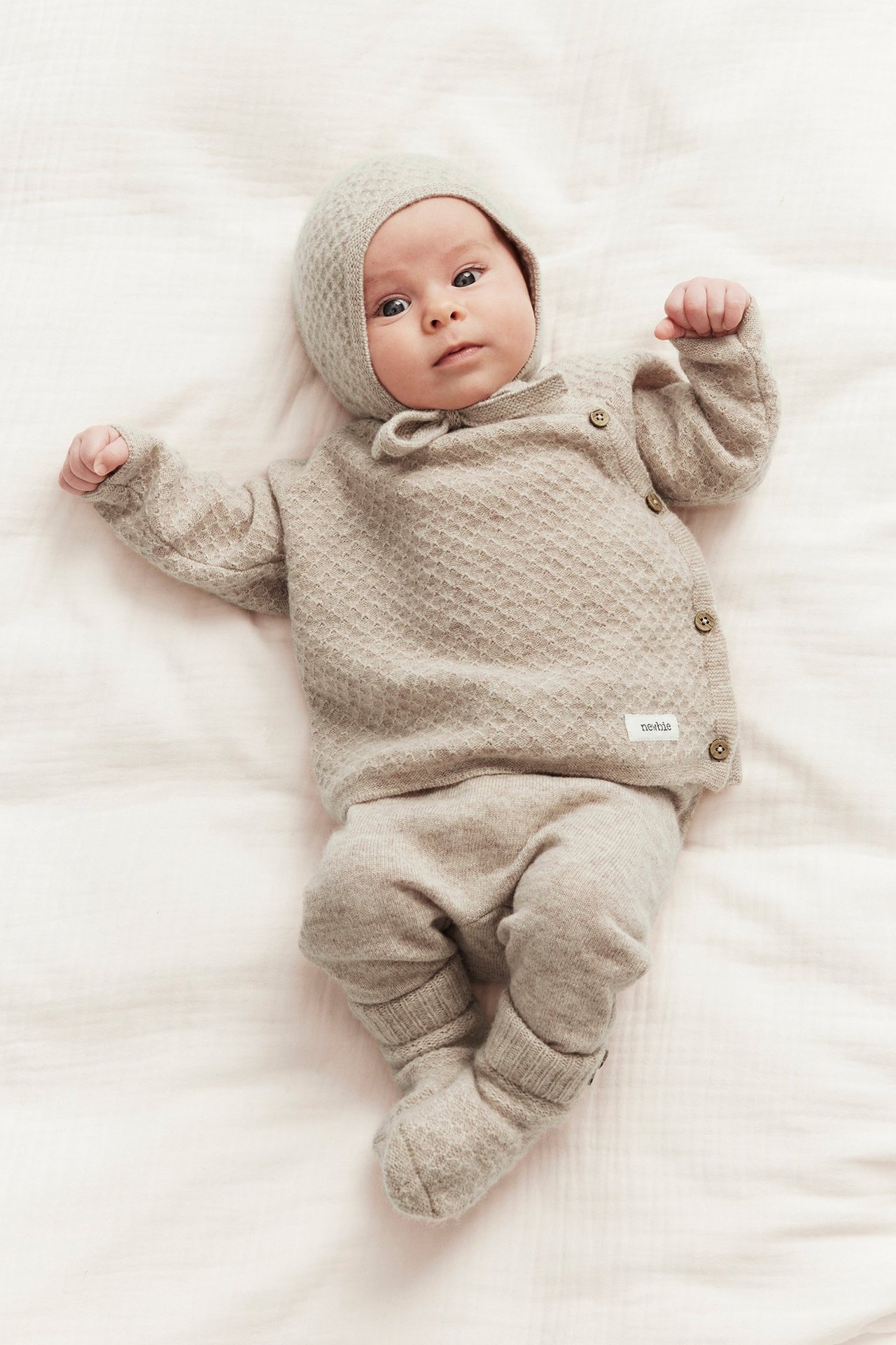 Vorderansicht: Baby in beiger melierter Strickjacke, passender Mütze und Hose.