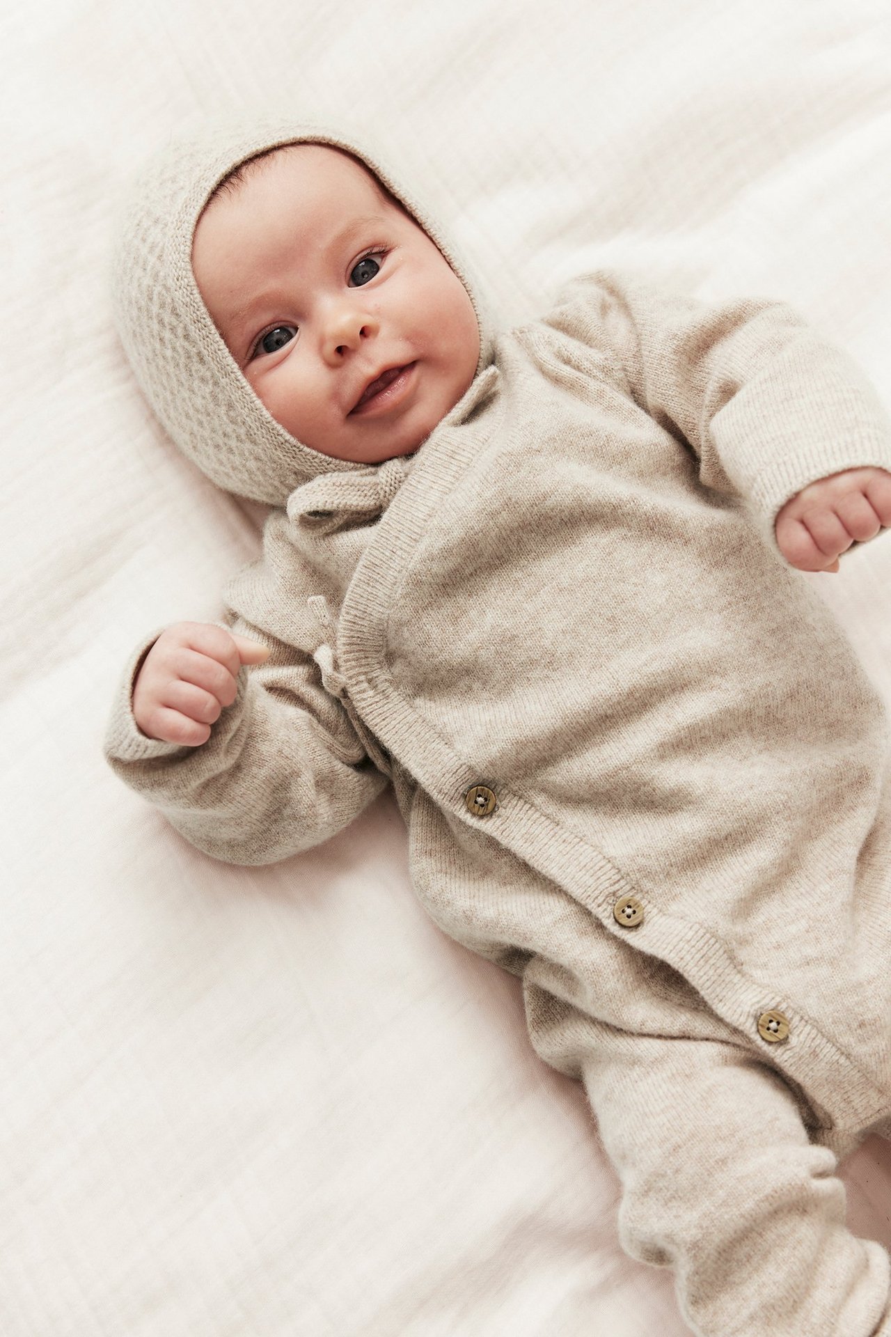 Top view of a baby wearing a beige melange wool-cashmere wrap jumpsuit with buttons and a matching knitted bonnet.