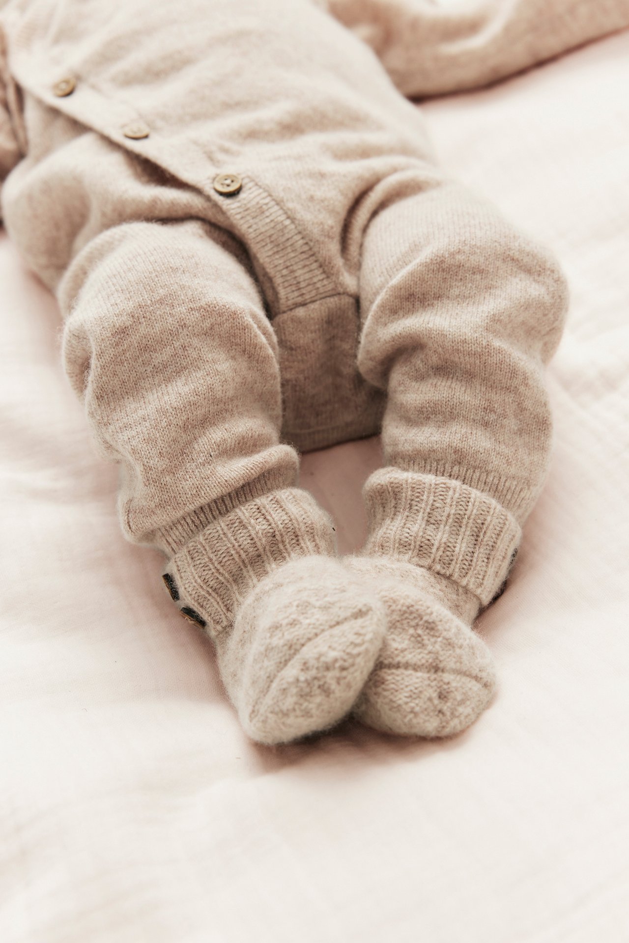 Nahaufnahme eines Babys in einem beigen melierten Wickel-Overall aus Wolle-Kaschmir mit Knöpfen und Rippbündchen.