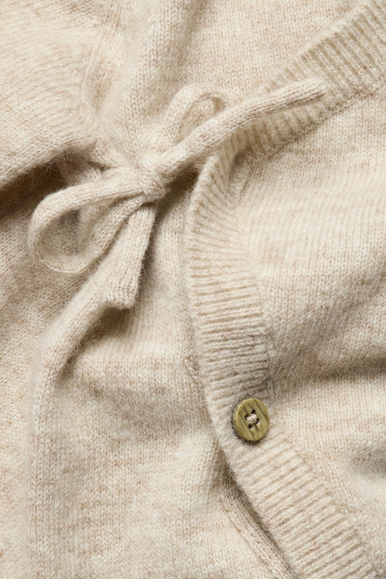 Nahaufnahme eines beigen melierten glatt gestrickten Baby-Wickeloveralls mit Schleife und Knopfverschluss vorne.