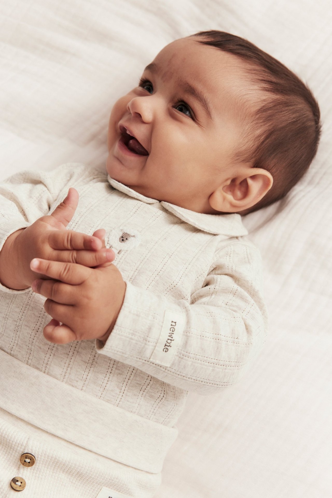 Close-up front view of a smiling baby in a beige melange pointelle rib long-sleeved body with collar and embroidery.