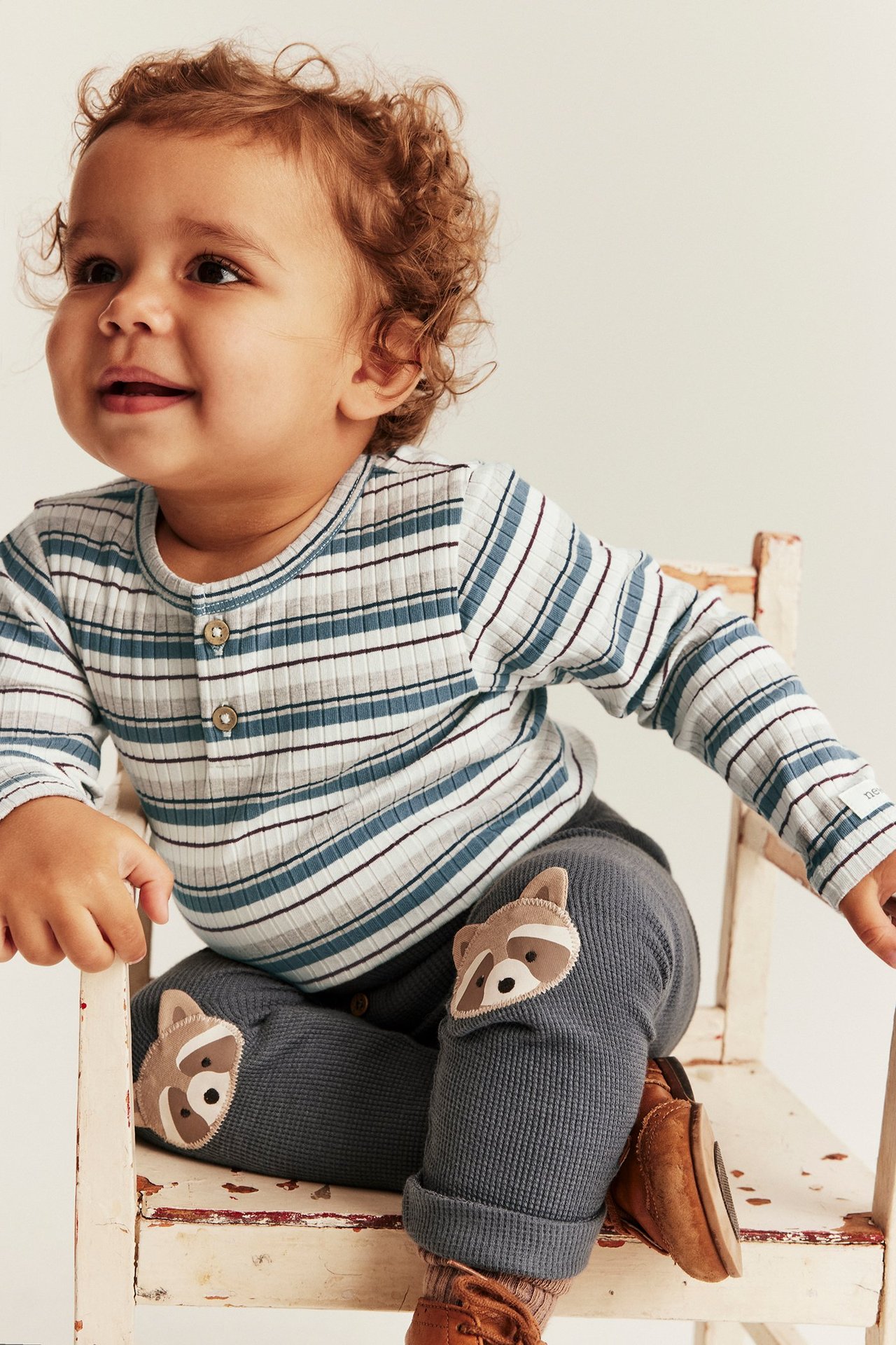 Front view: Baby wears a blue, grey striped ribbed bodysuit, front buttons, and dark grey raccoon knee pants.