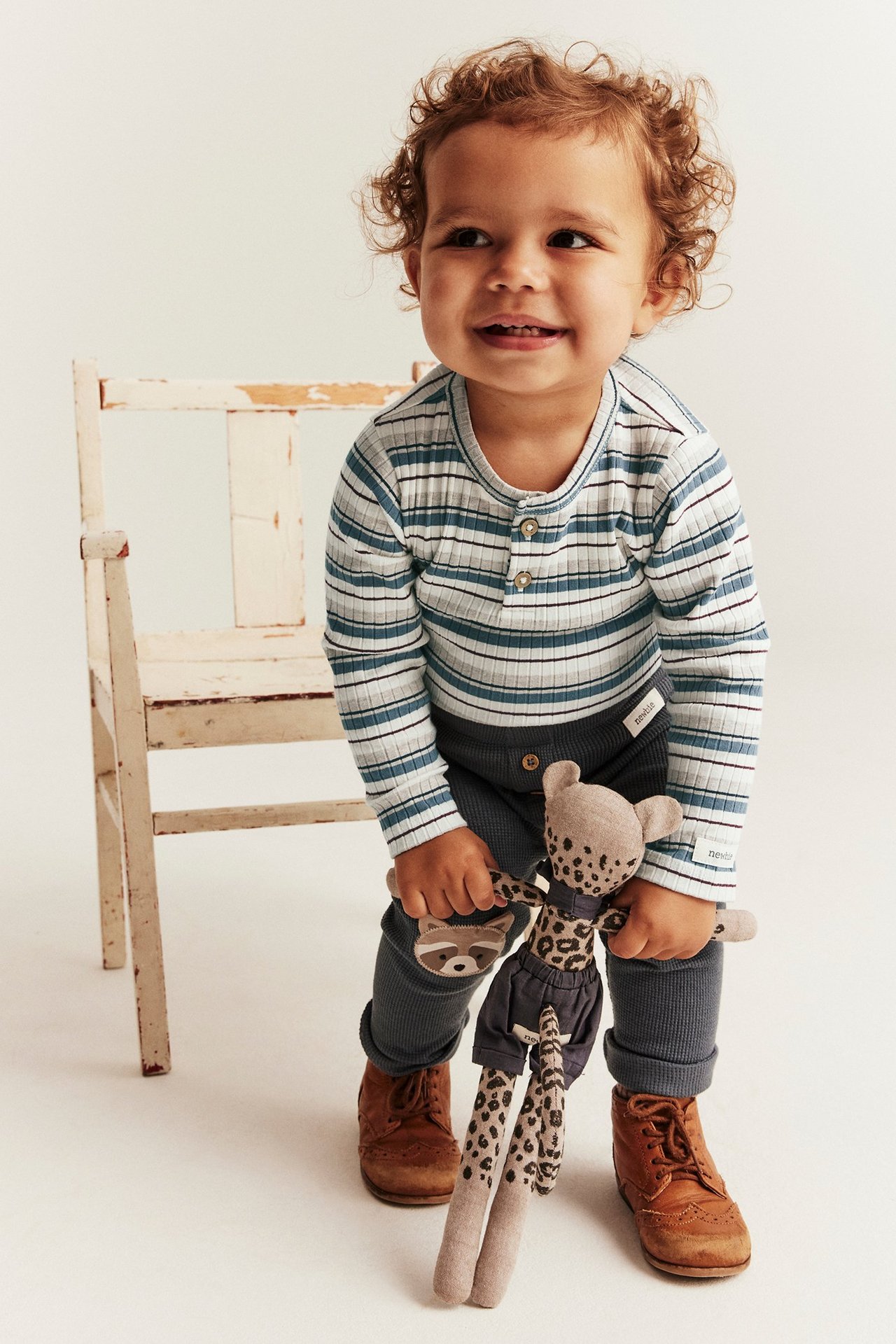 Baby in einem blau gestreiften, gerippten Langarm-Body mit Knöpfen, dunkelblauen Leggings und braunen Schuhen.