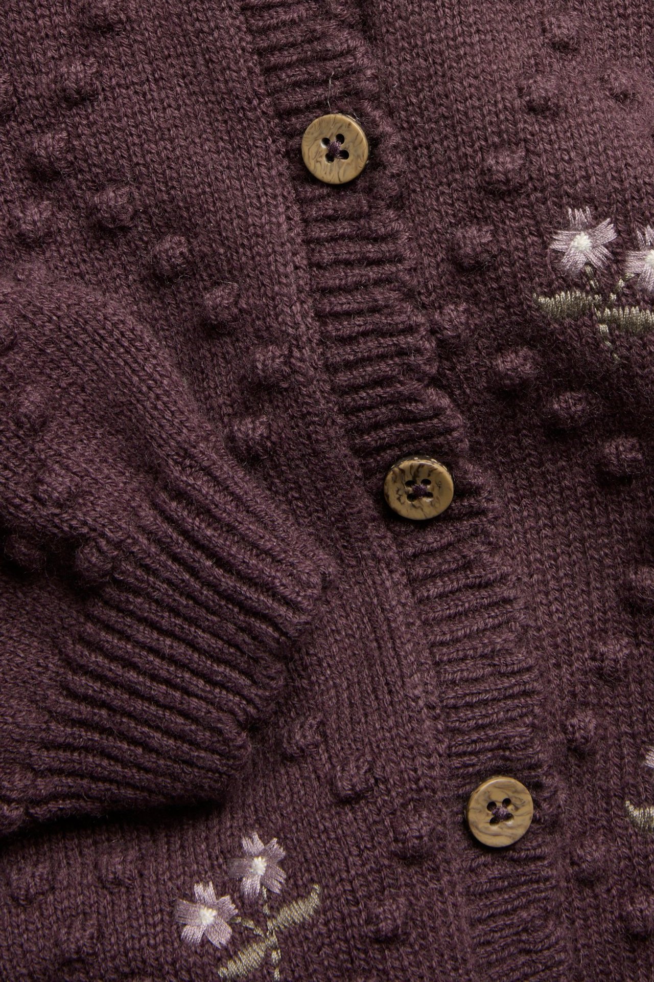 Nahaufnahme einer lila Wollmisch-Strickjacke mit Strickpunkten, gestickten Blumen und Holzknöpfen.