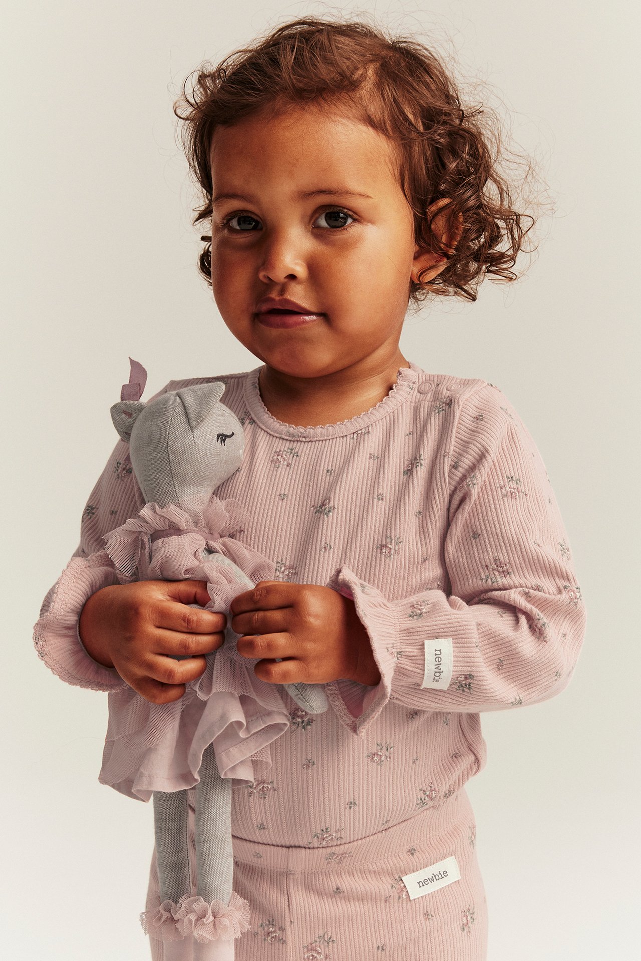 Vorderansicht eines Baby-Mädchens in rosa geripptem Blumen-Langarmbody und passender Hose, hält eine graue Puppe.