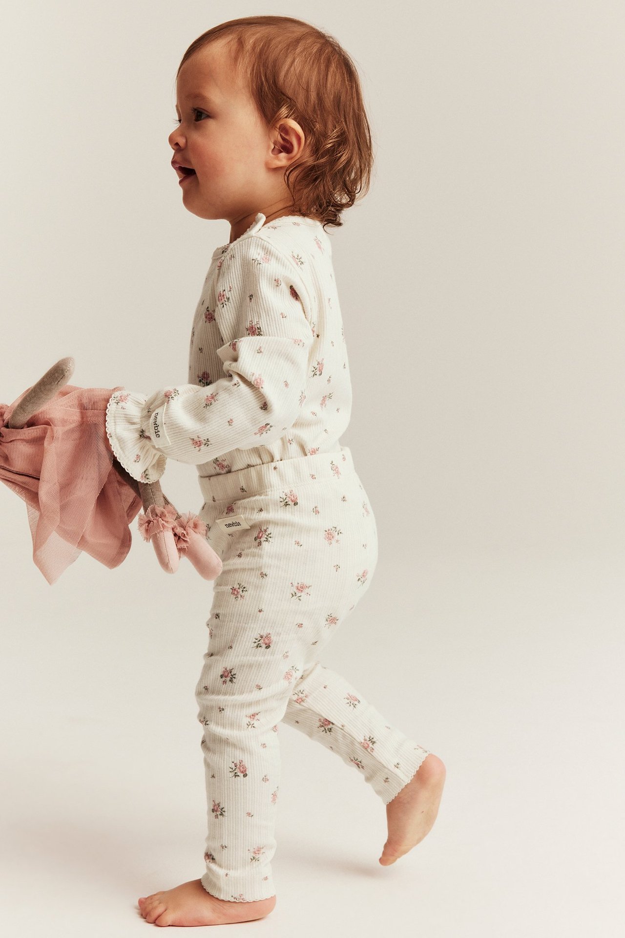 Seitenansicht eines Babys in cremeweißen gerippten Blumen-Leggings und passendem Oberteil, das eine rosa Puppe hält.