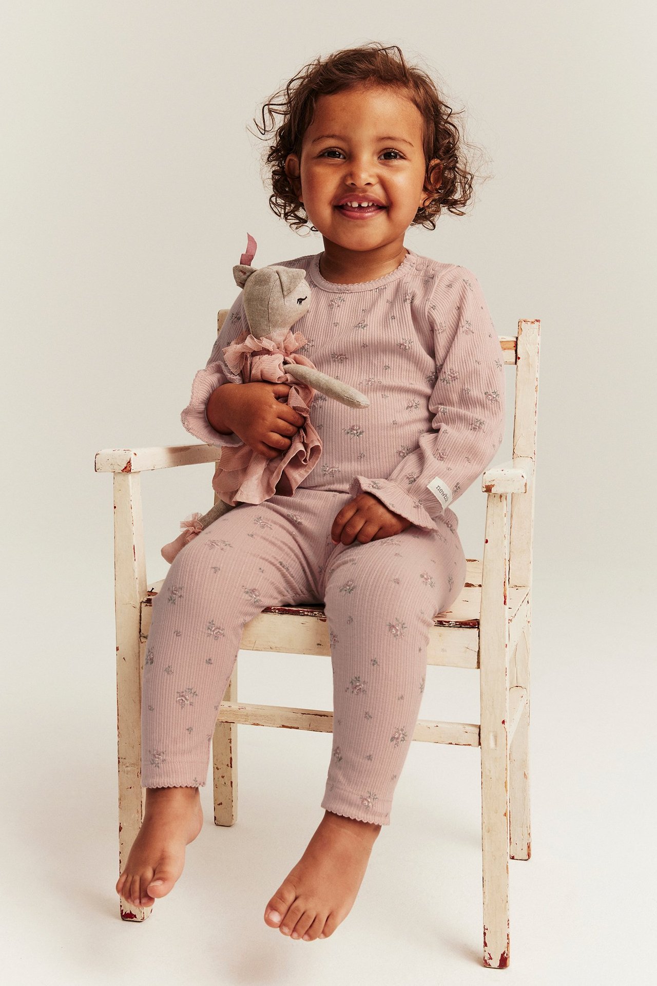 Smiling kid in pink ribbed floral leggings and matching top, holding a doll. Front view.
