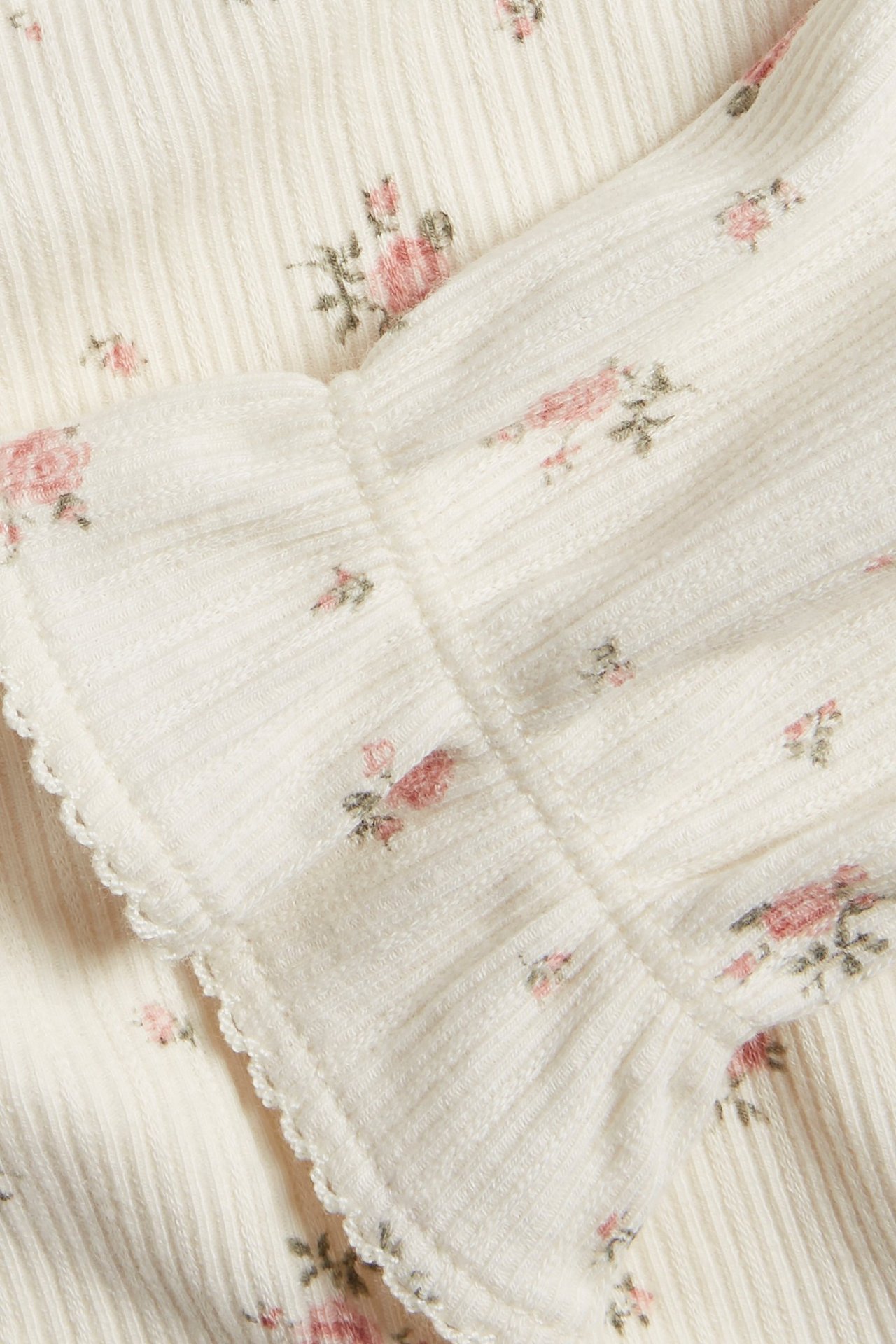 Nahaufnahme eines cremefarbenen gerippten Oberteils mit kleinem rosa Blumenmuster und Picot-Rüsche.