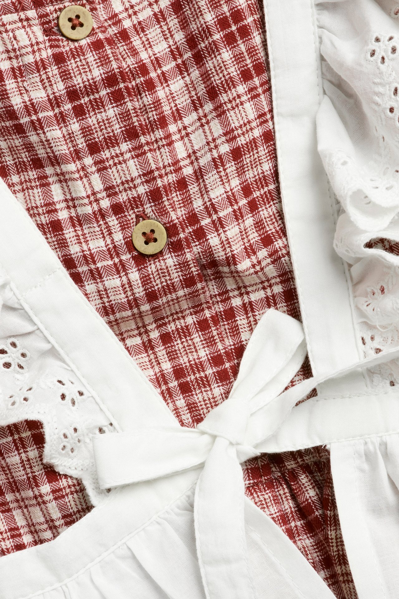 Nahaufnahme: Rot kariertes Baumwollkleid mit Holzknöpfen, weißer bestickter Schürze mit Schleife.