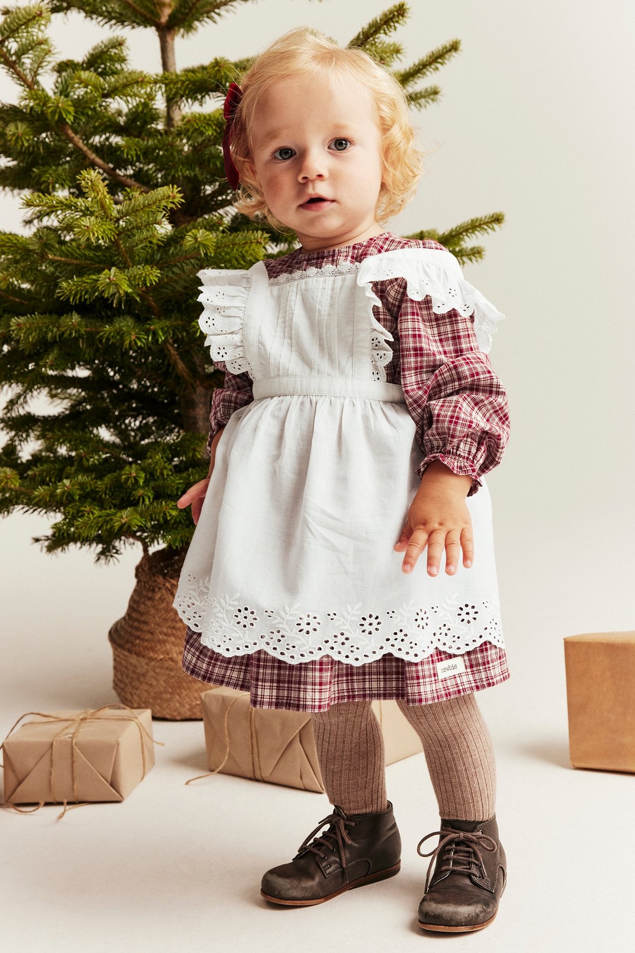 Front view of baby in red checkered dress, white eyelet apron, brown tights, and dark boots.