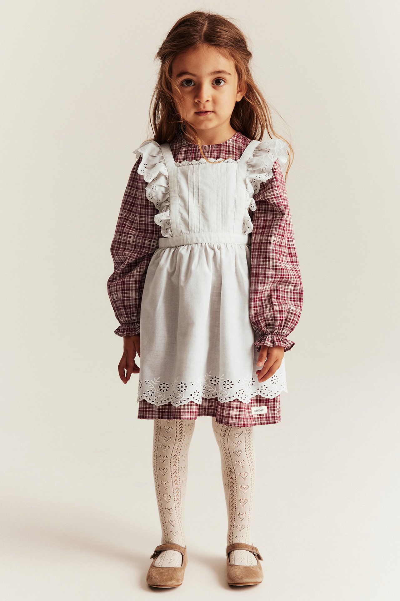 Kid wears red checkered dress, white ruffled apron, light tights, brown shoes. Front view.