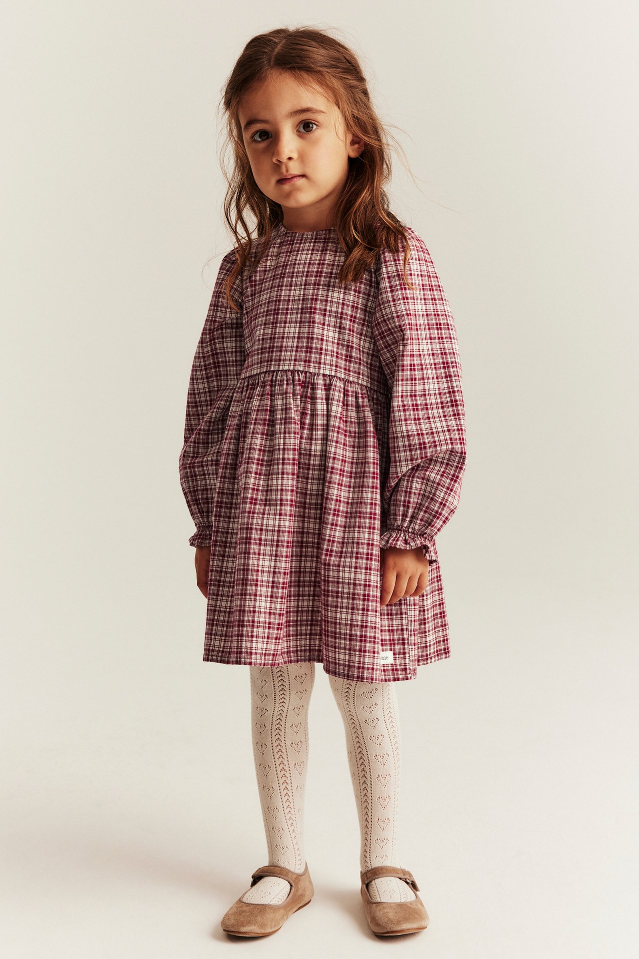 Front view: kid in red checked cotton dress, white patterned tights, and brown Mary Jane shoes.