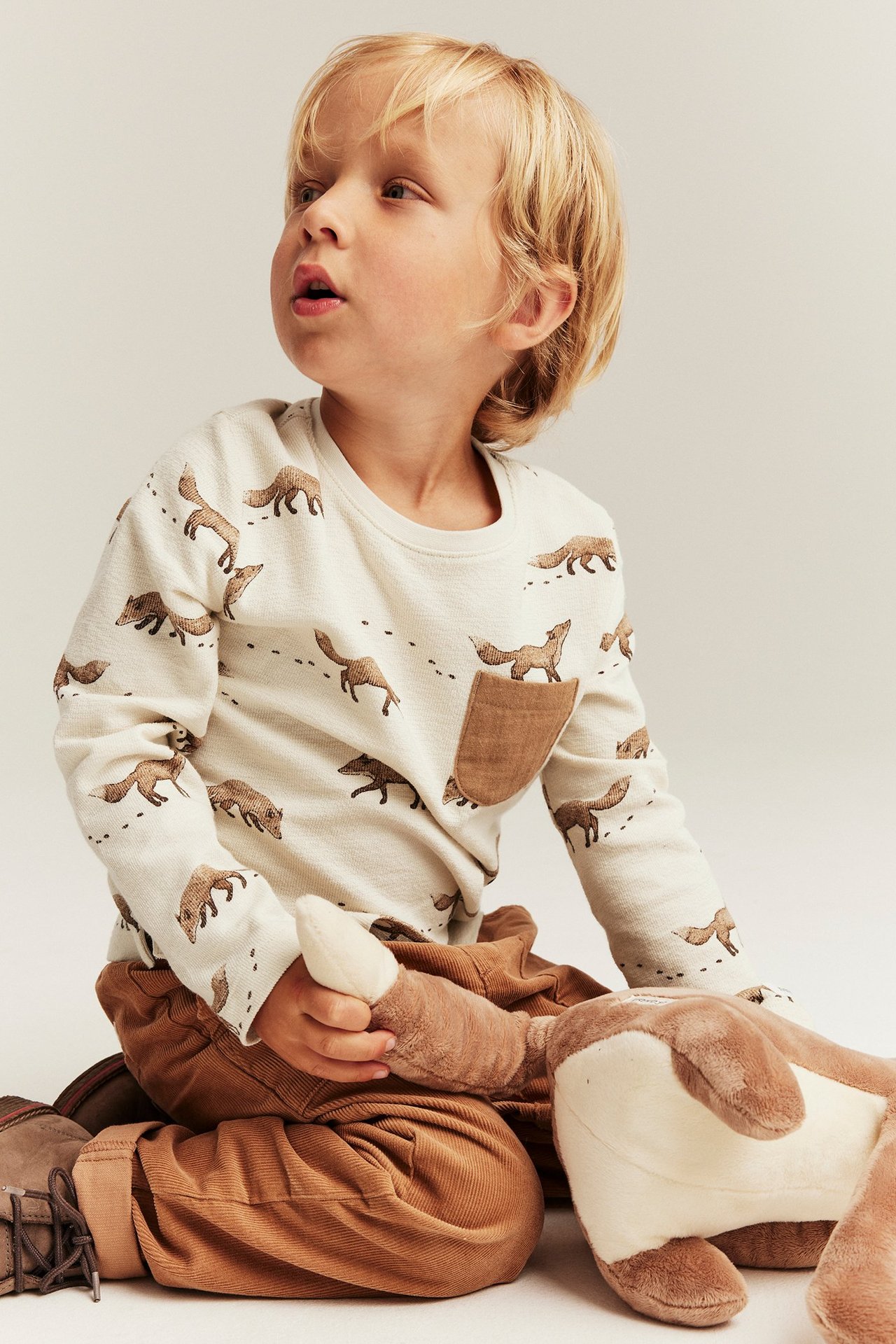 Kind in beigem Langarmshirt mit Fuchsmuster, brauner Tasche, brauner Hose, hält ein Plüschtier.