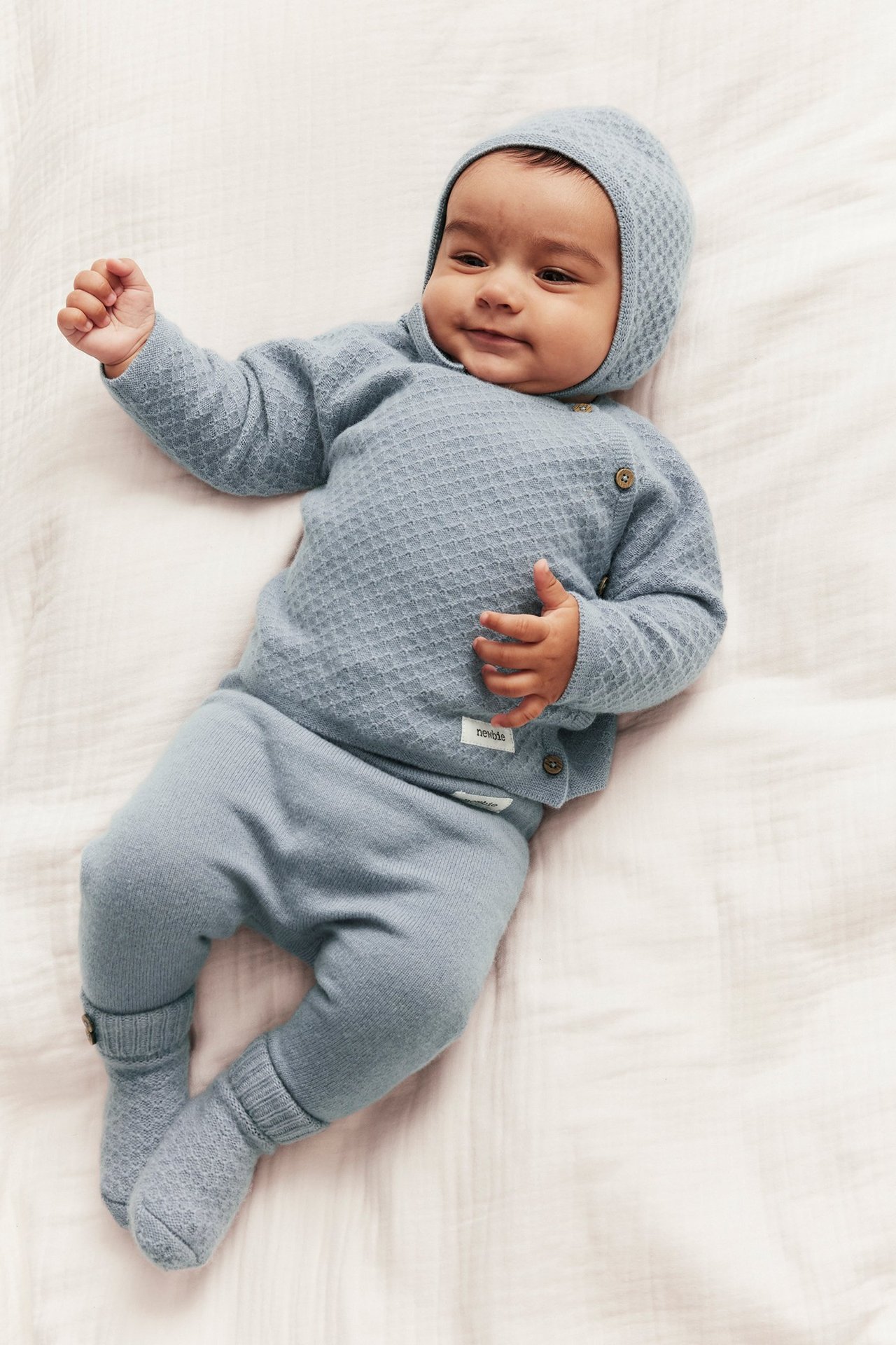 Top view of baby in light blue plain knit pants, textured sweater with buttons, and bonnet.