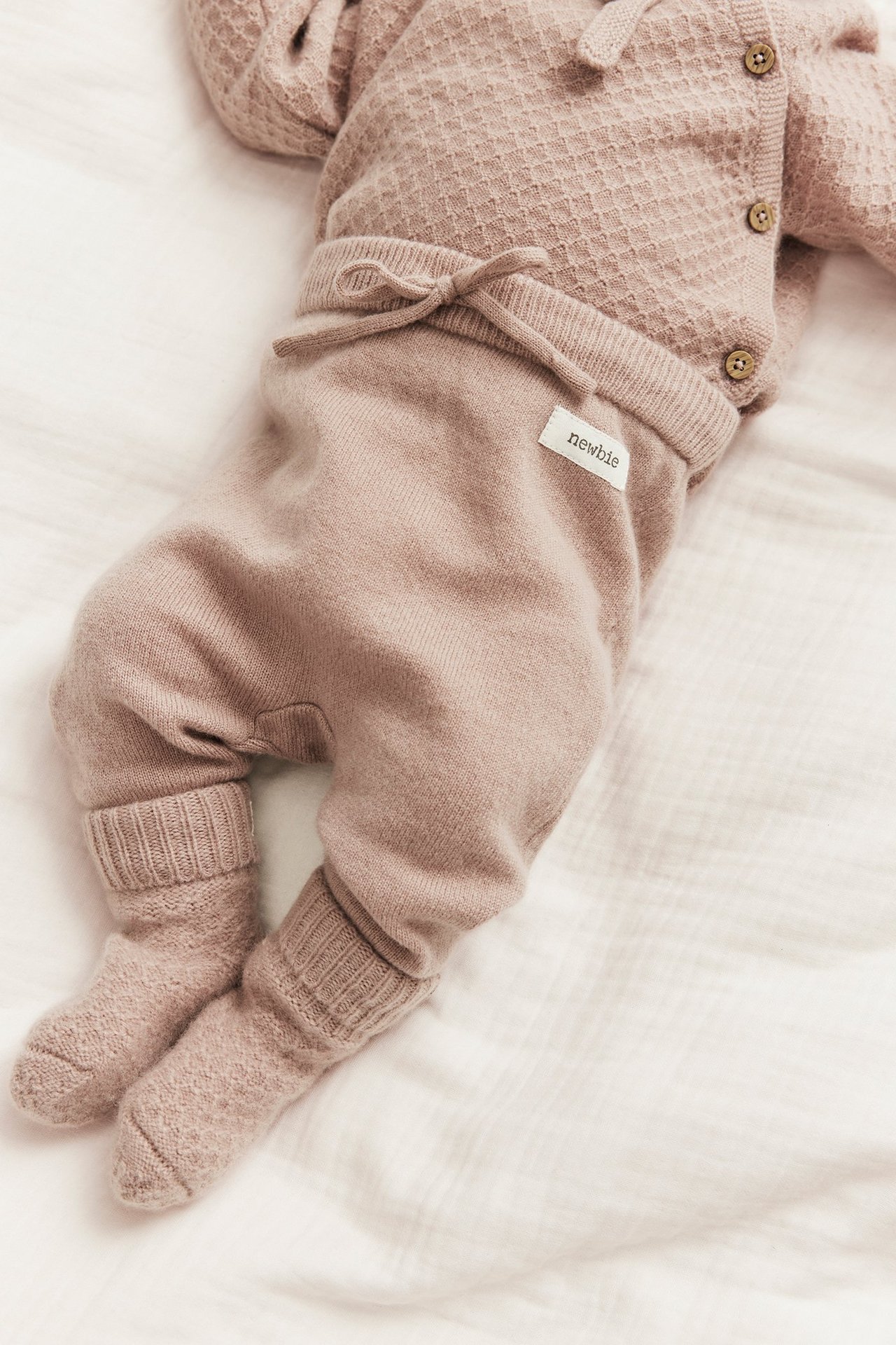 Close-up of a baby wearing pink smooth-knitted pants with a drawstring, a textured cardigan, and knitted socks.