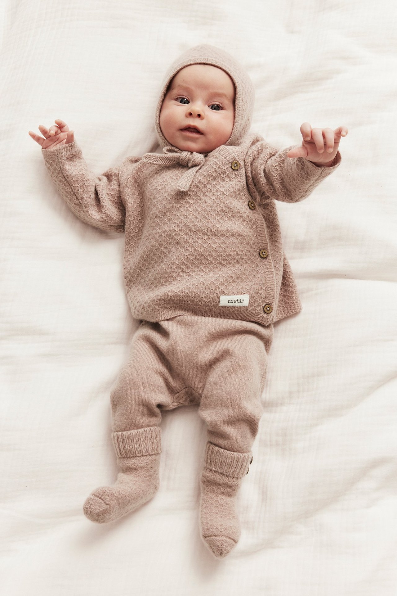 Front view of a baby wearing pink wool-cashmere blend pants, a textured knit wrap cardigan, bonnet, and socks.