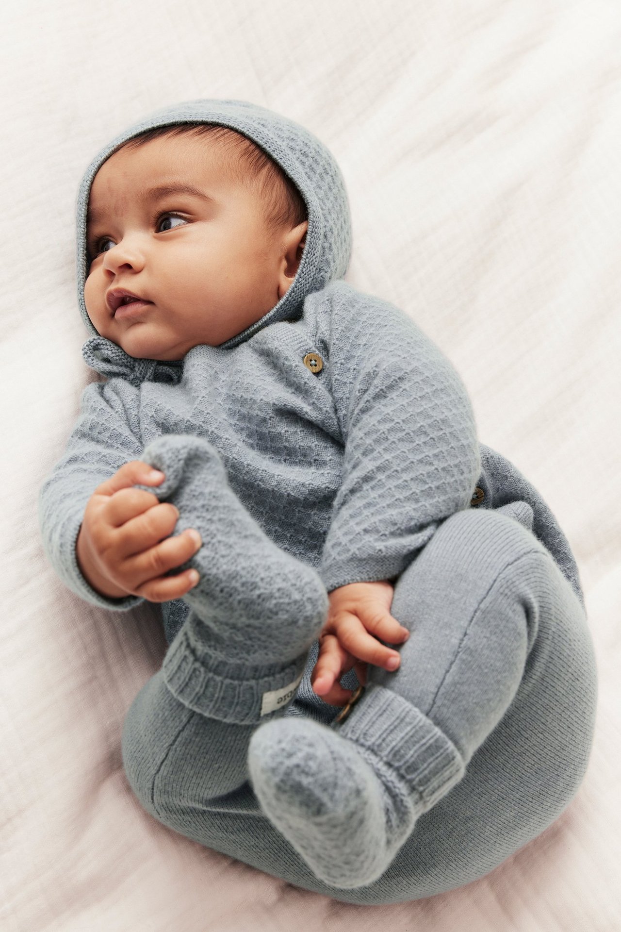 Aufnahme von oben: Baby in hellblauer strukturierter Strick-Woll-Kaschmir-Strickjacke, passender Hose, Mütze und Schühchen.