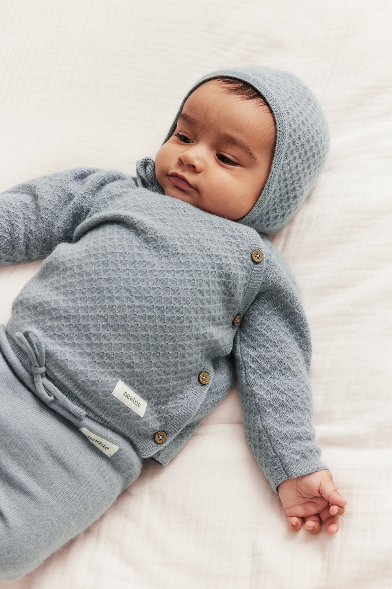 Top view of a baby wearing a light blue structured knit wool-cashmere cardigan with side buttons, matching bonnet, and trousers.