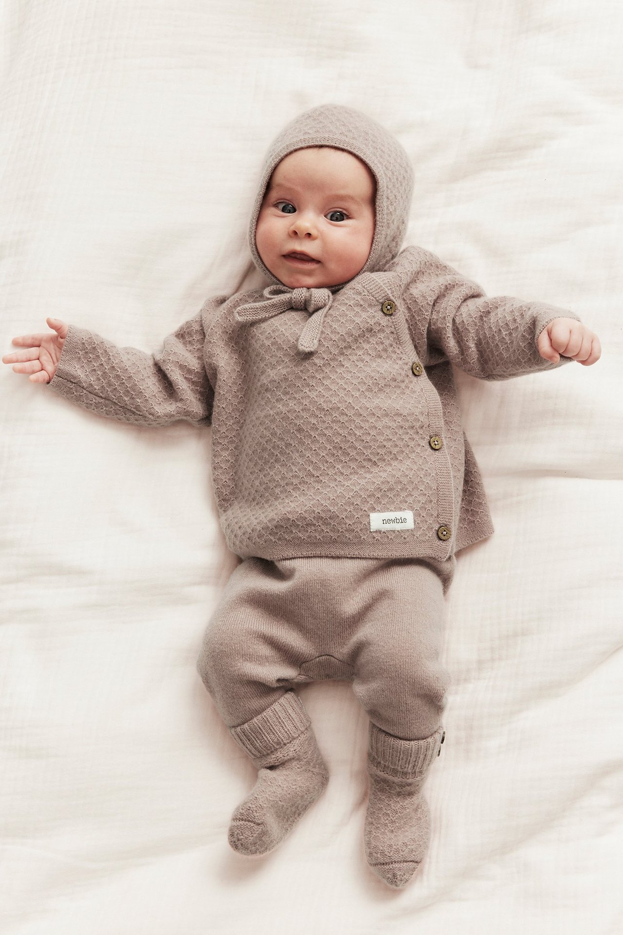 Front view: baby in a muted pink textured knit wool-cashmere cardigan with side buttons, matching bonnet, trousers, and socks.