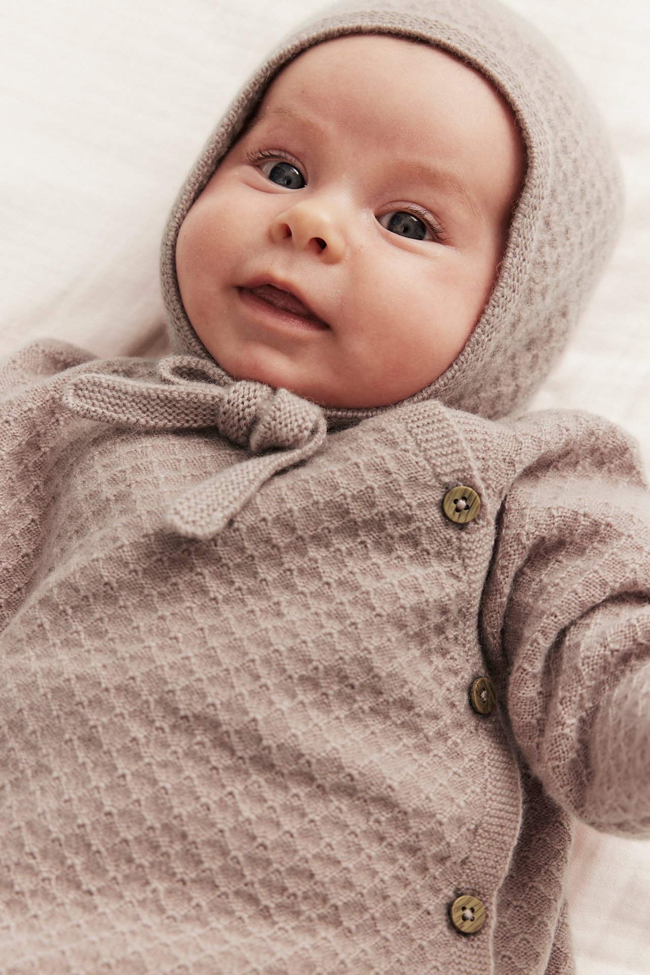Close-up of a baby wearing a light pink structured knit wool-cashmere cardigan with side buttons and a matching bonnet.
