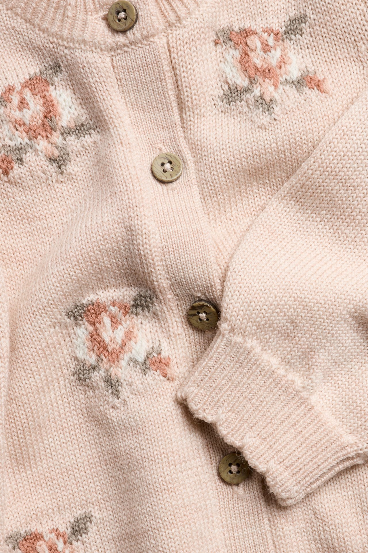 Nahaufnahme: rosa Strick-Wolljacke mit Jacquard-Rosen, braunen Knöpfen, gerippten Wellenkanten.