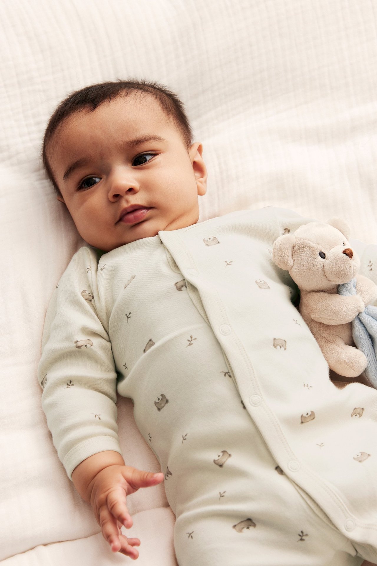 Close-up of a baby in light beige cotton pyjamas with bear print, snap buttons, next to a teddy bear.