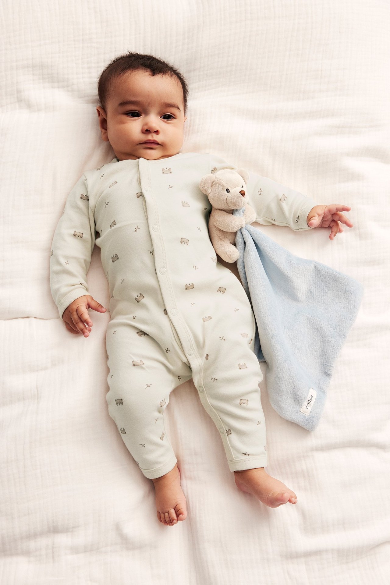 Baby in light beige cotton pyjamas with teddy bear print, holding a teddy bear and blue blanket.
