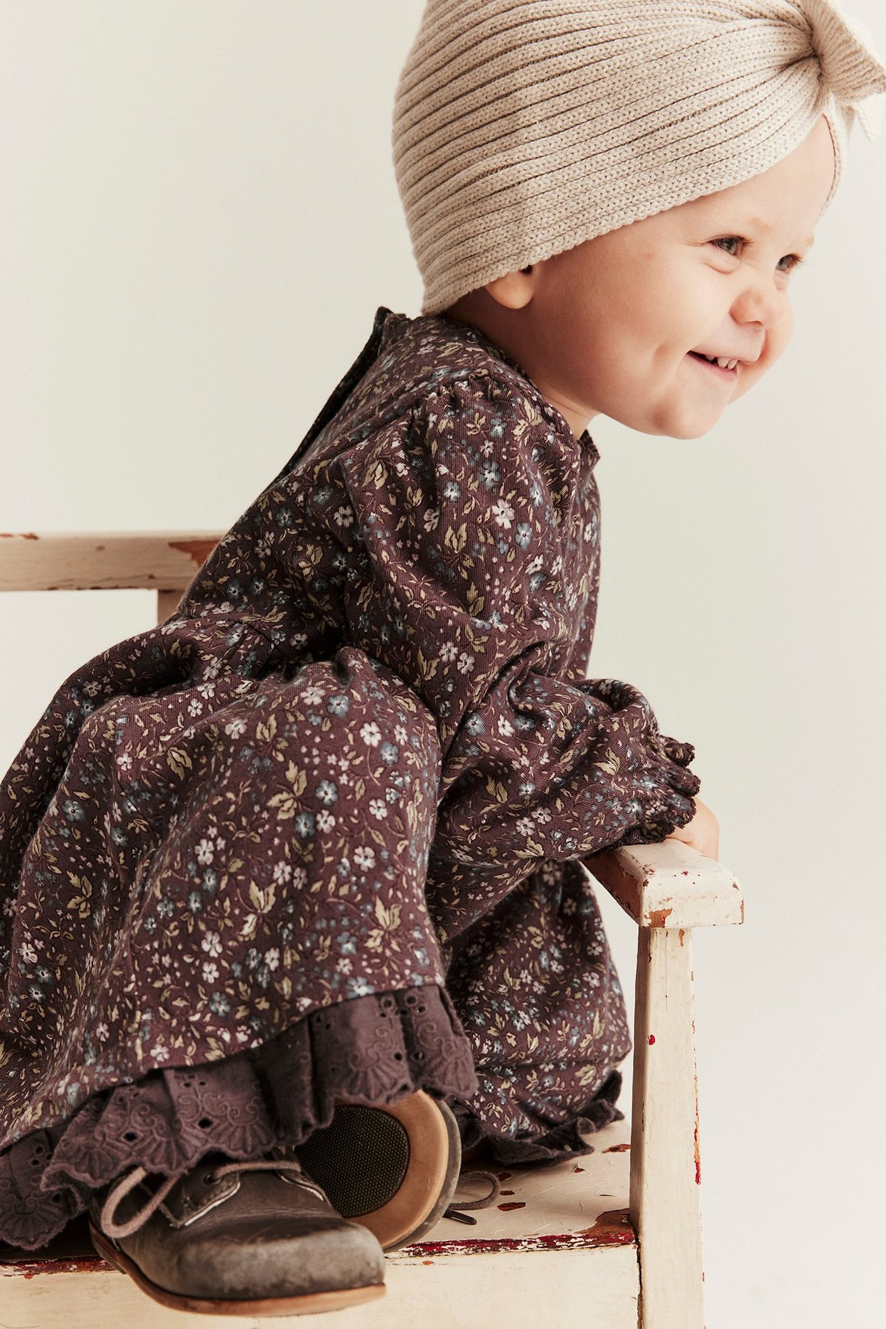 Seitenansicht eines Babys in einem dunkelvioletten geblümten Jerseykleid mit Spitzenbesatz, beigem Strickturban und braunen Schuhen.