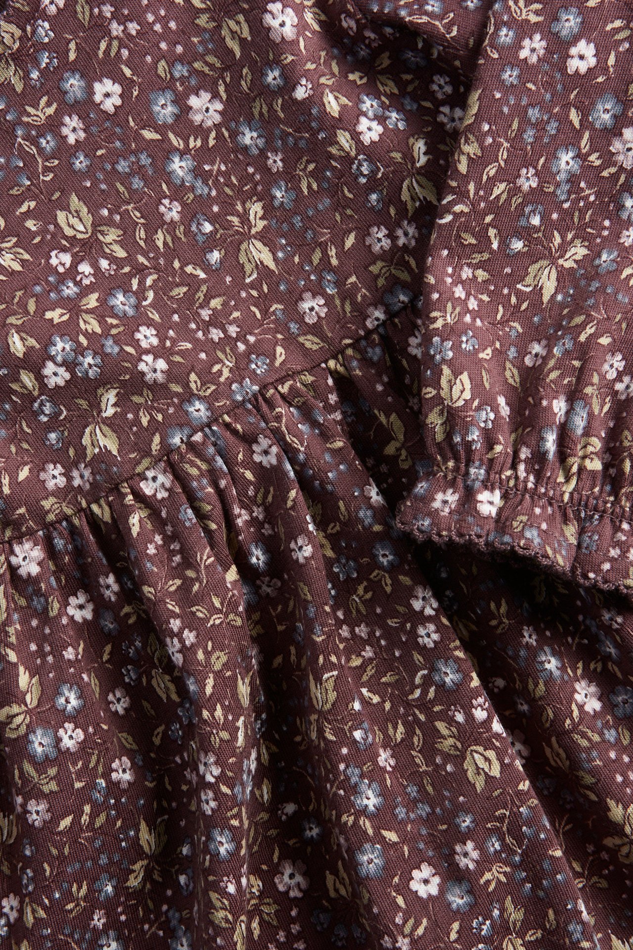 Nahaufnahme eines lila geblümten Jerseykleides mit kleinen weißen und blauen Blumen, zeigt Rüschen und elastischen Ärmelbund.