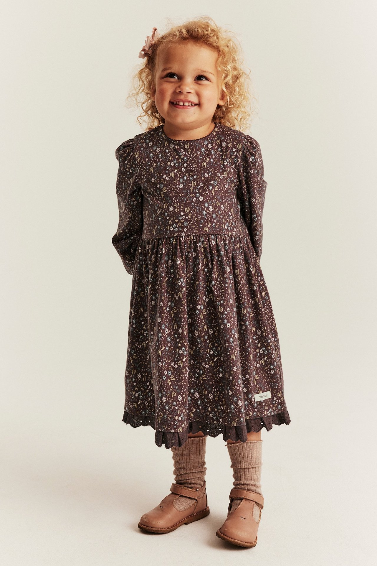 Front view: A smiling kid wears a dark floral jersey dress, light brown ribbed socks, and brown Mary Jane shoes.