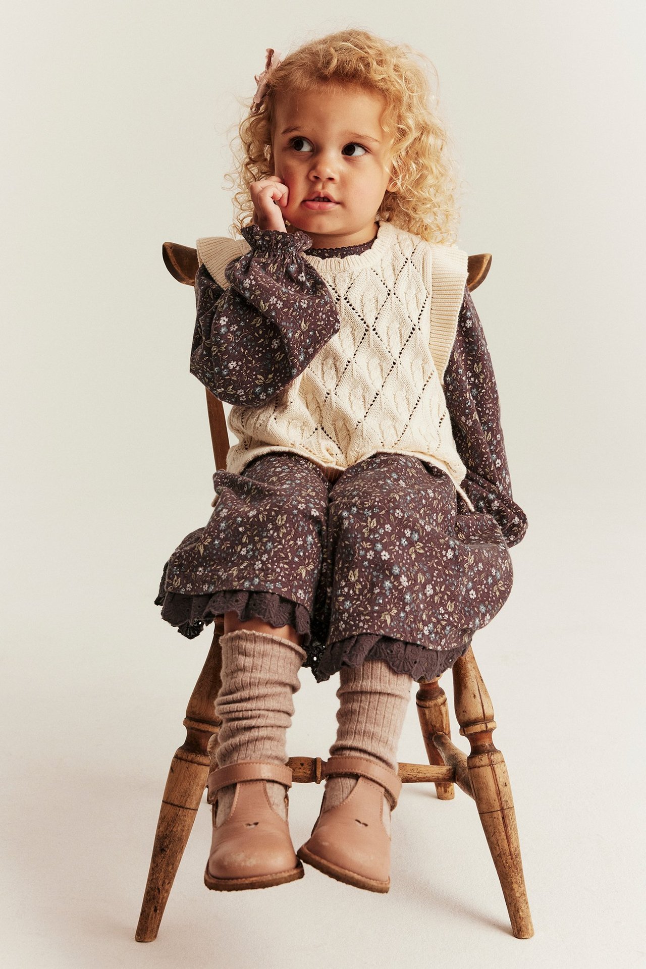 Front view: Kid in a dark floral jersey dress, cream knitted vest, beige leg warmers, and brown shoes.