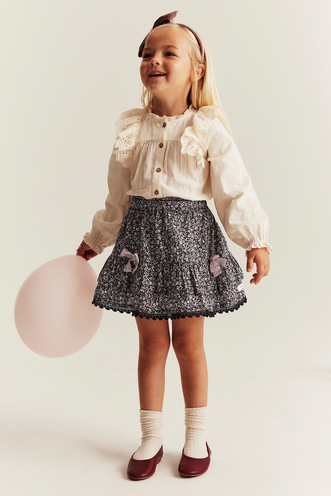 Front view: Kid wears a black floral tiered skirt with bows, lace trim, cream blouse, and red shoes.