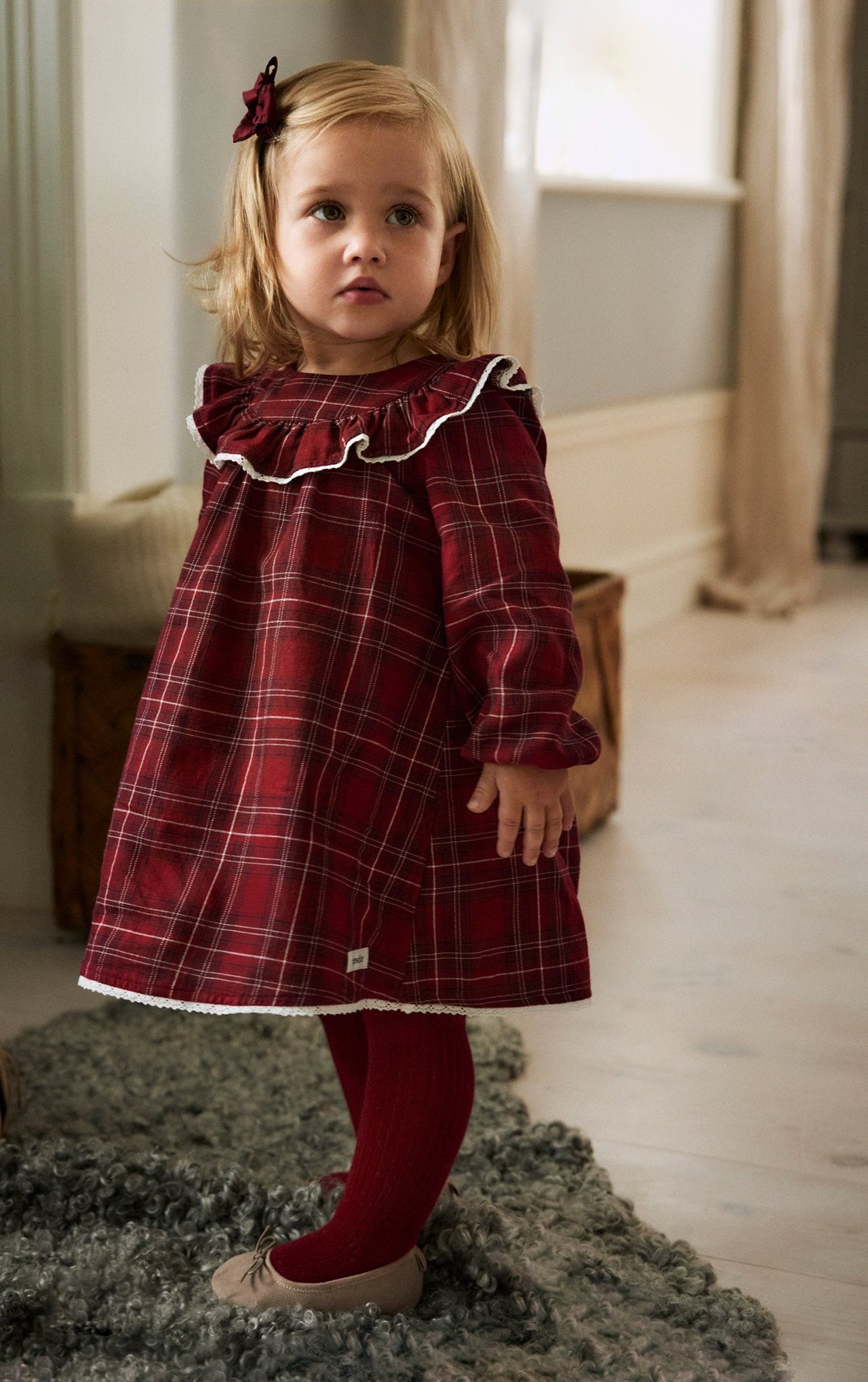 Front view: baby in a red checkered cotton dress with white ruffles, red tights, beige shoes, red bow.