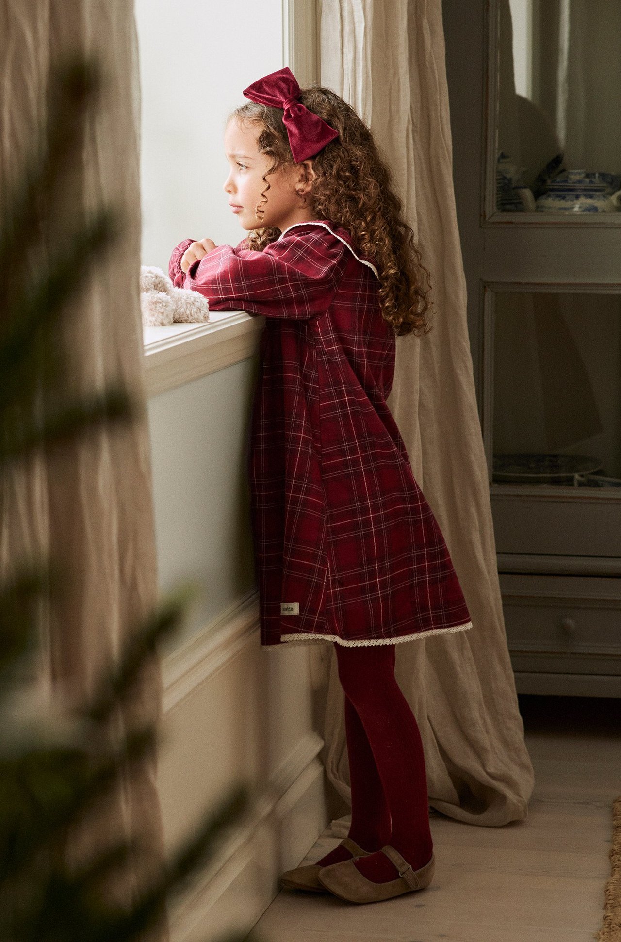Kid's side view, red plaid cotton dress with white collar and lace, red velvet bow, tights, and beige shoes.