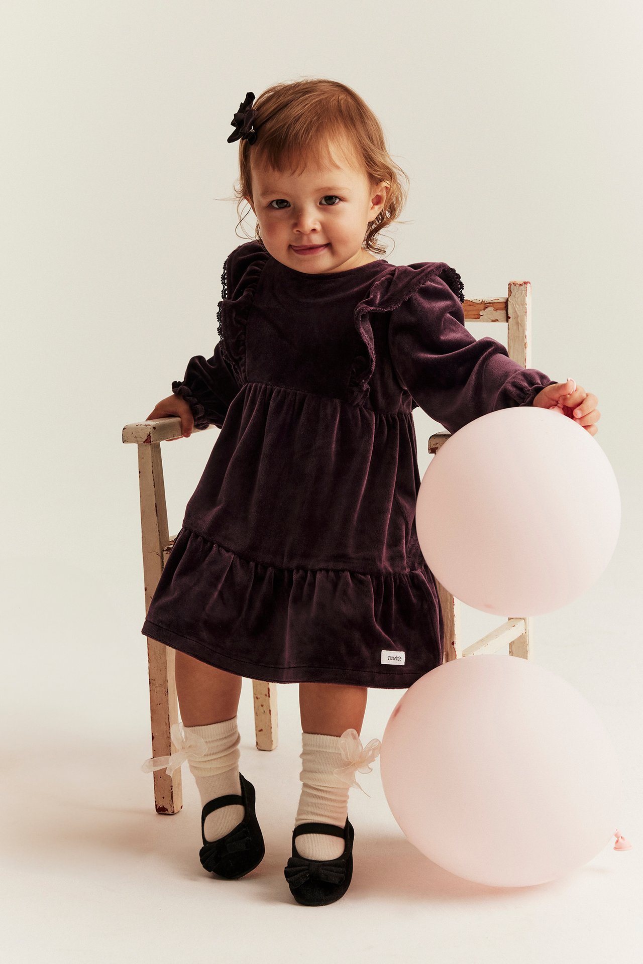 Front view of a baby wearing a lilac velour dress with ruffles, black Mary Jane shoes, and cream socks.