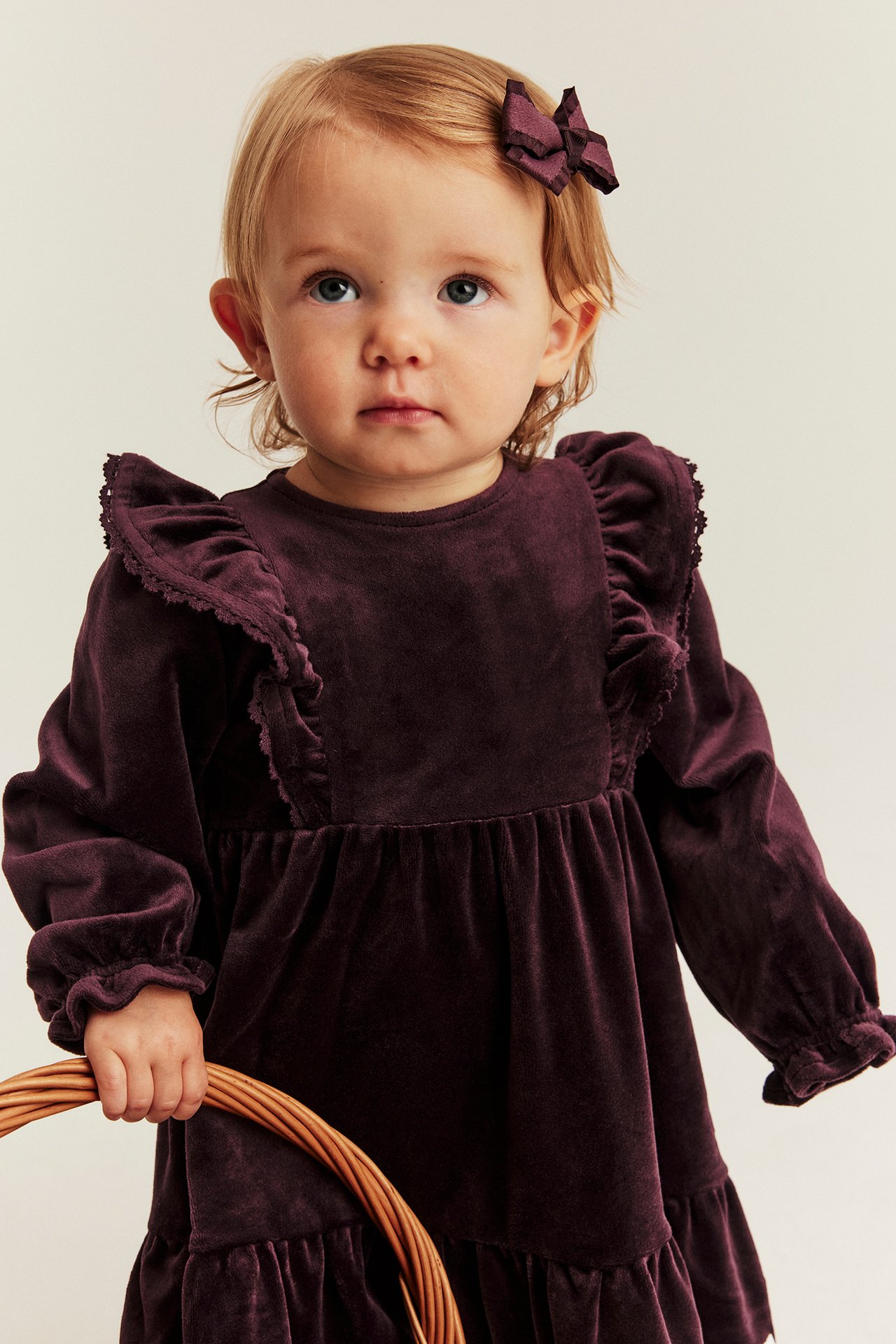 Vorderansicht eines Baby-Mädchens in dunkelviolettem Samtkleid mit Rüschen und passender Schleife, hält einen Weidenkorb.