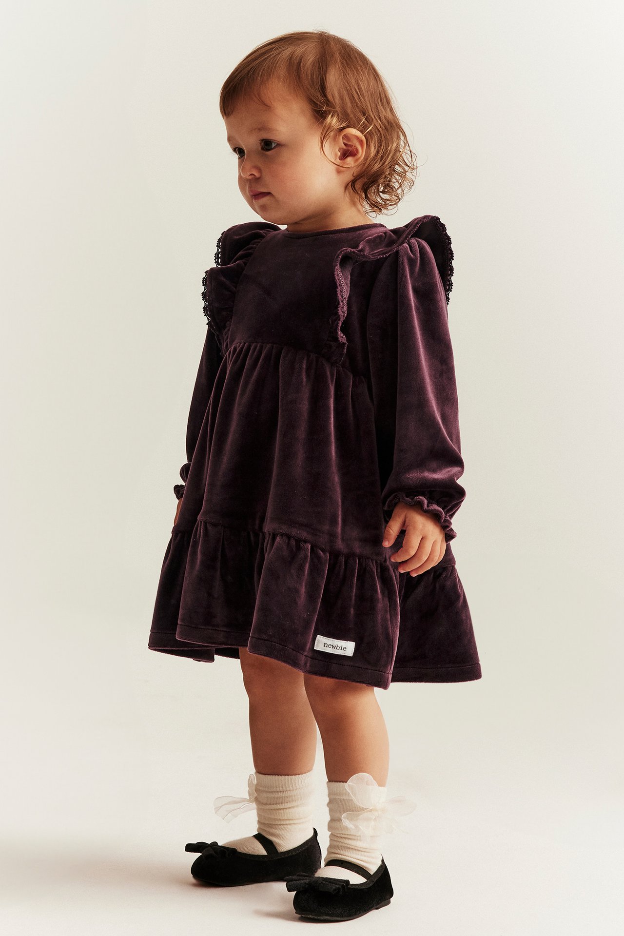 Side view of a baby in a lilac velour dress with shoulder and hem ruffles, cream socks, and black shoes.