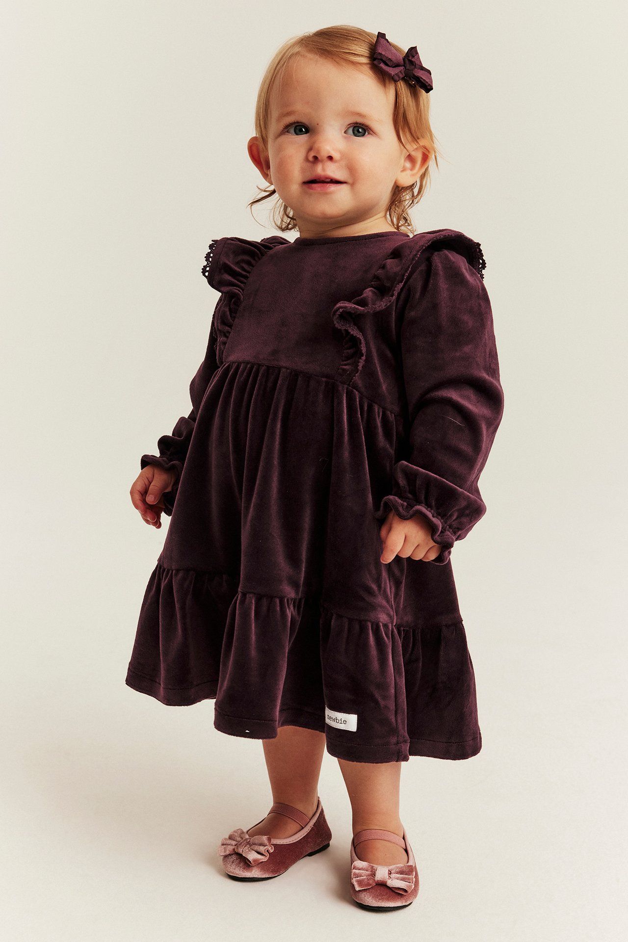 Vorderansicht eines Babys in einem dunkelvioletten Samtkleid, passender Schleife und rosa Samtschuhen.