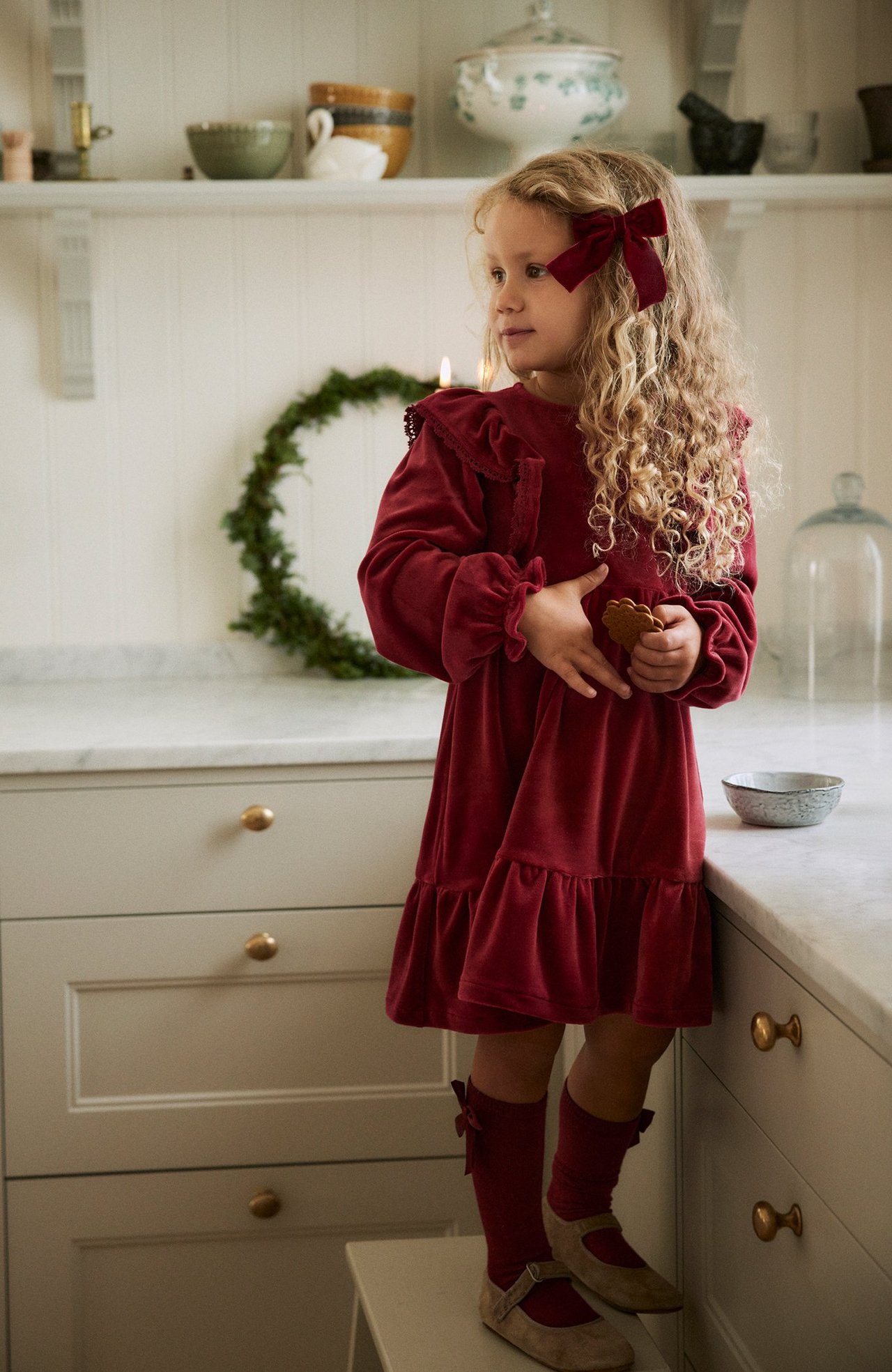 Full shot: Kid in red velour ruffle dress, red socks, beige shoes, and red hair bow, holding a cookie.