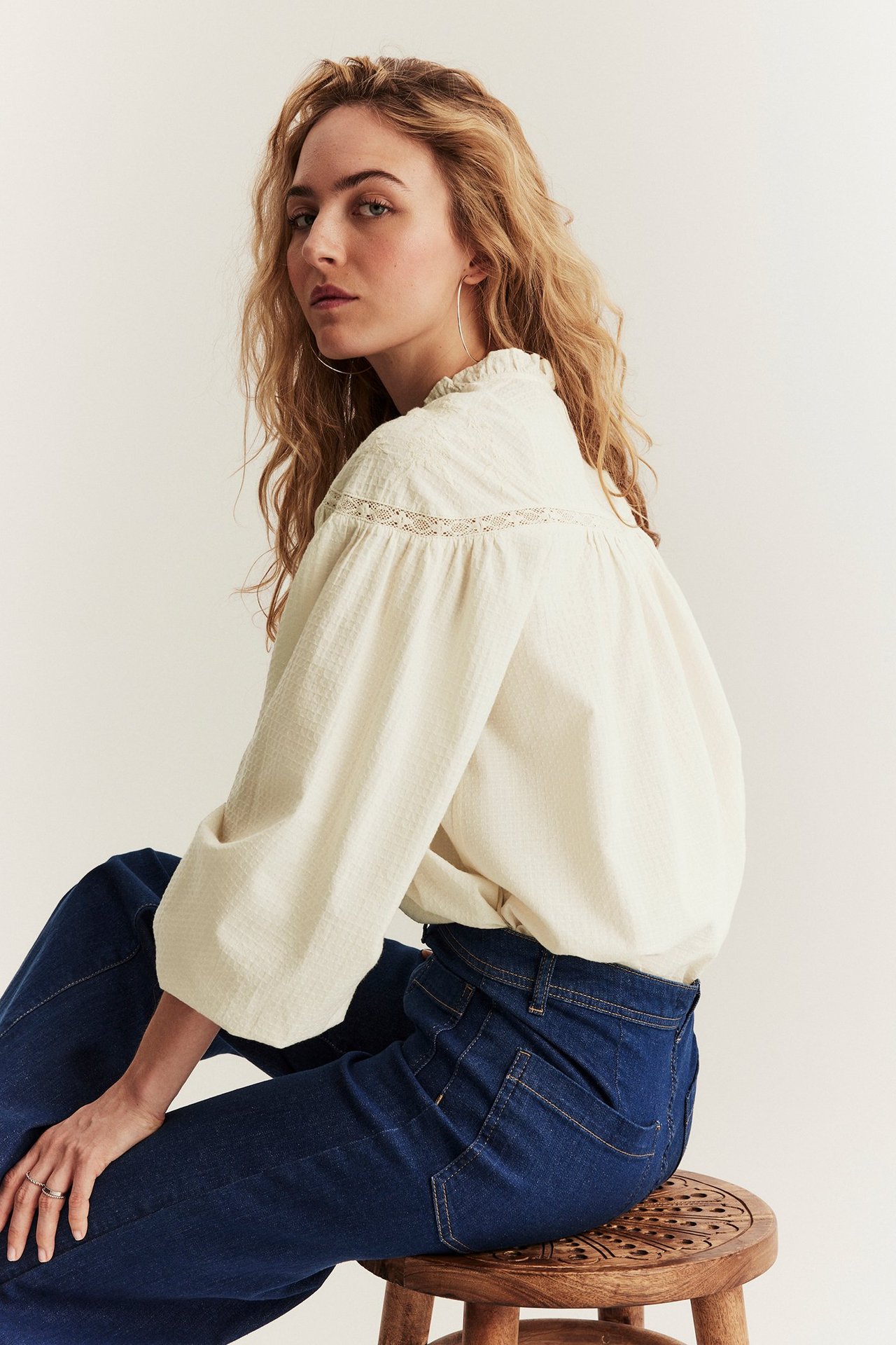 Seitenansicht einer Frau in einer hellbeigen Baumwollbluse mit Spitze und dunkelblauen Jeans.