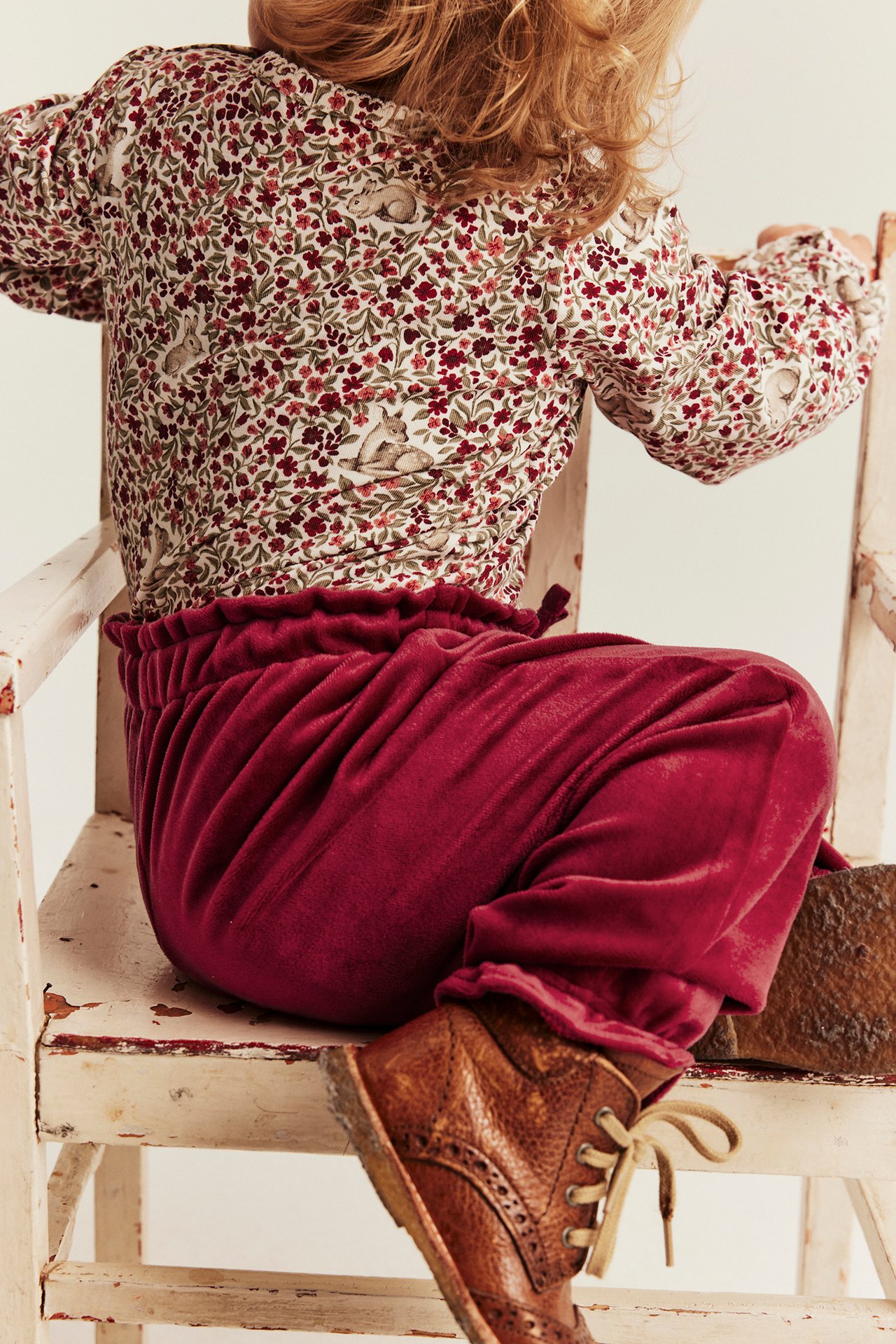 Back view kid in dark red velvet drawstring pants, cream floral bunny long-sleeve top, brown lace-up boots.