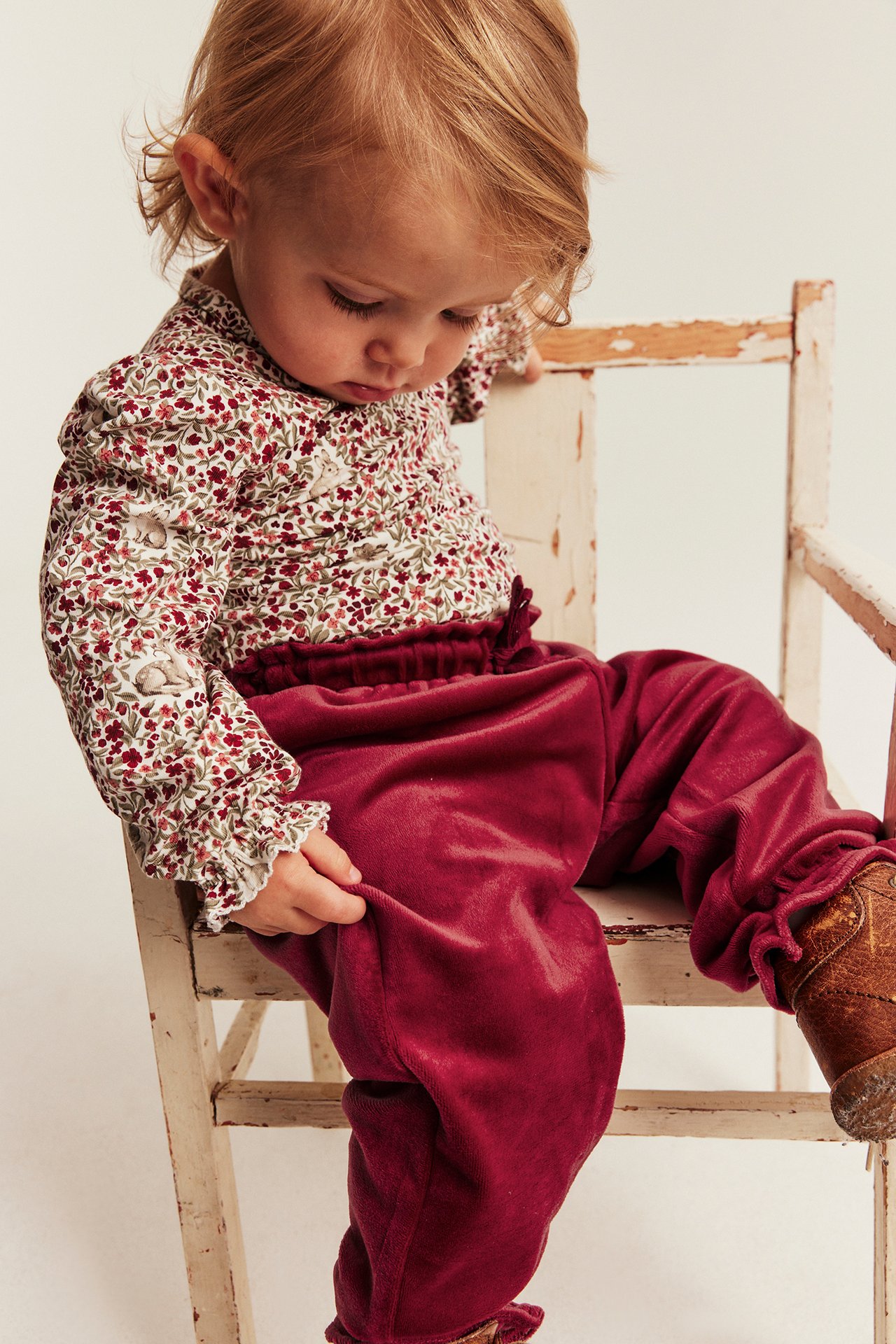 Close-up of baby wearing maroon velvet trousers, a white floral long-sleeve top, and brown shoes.