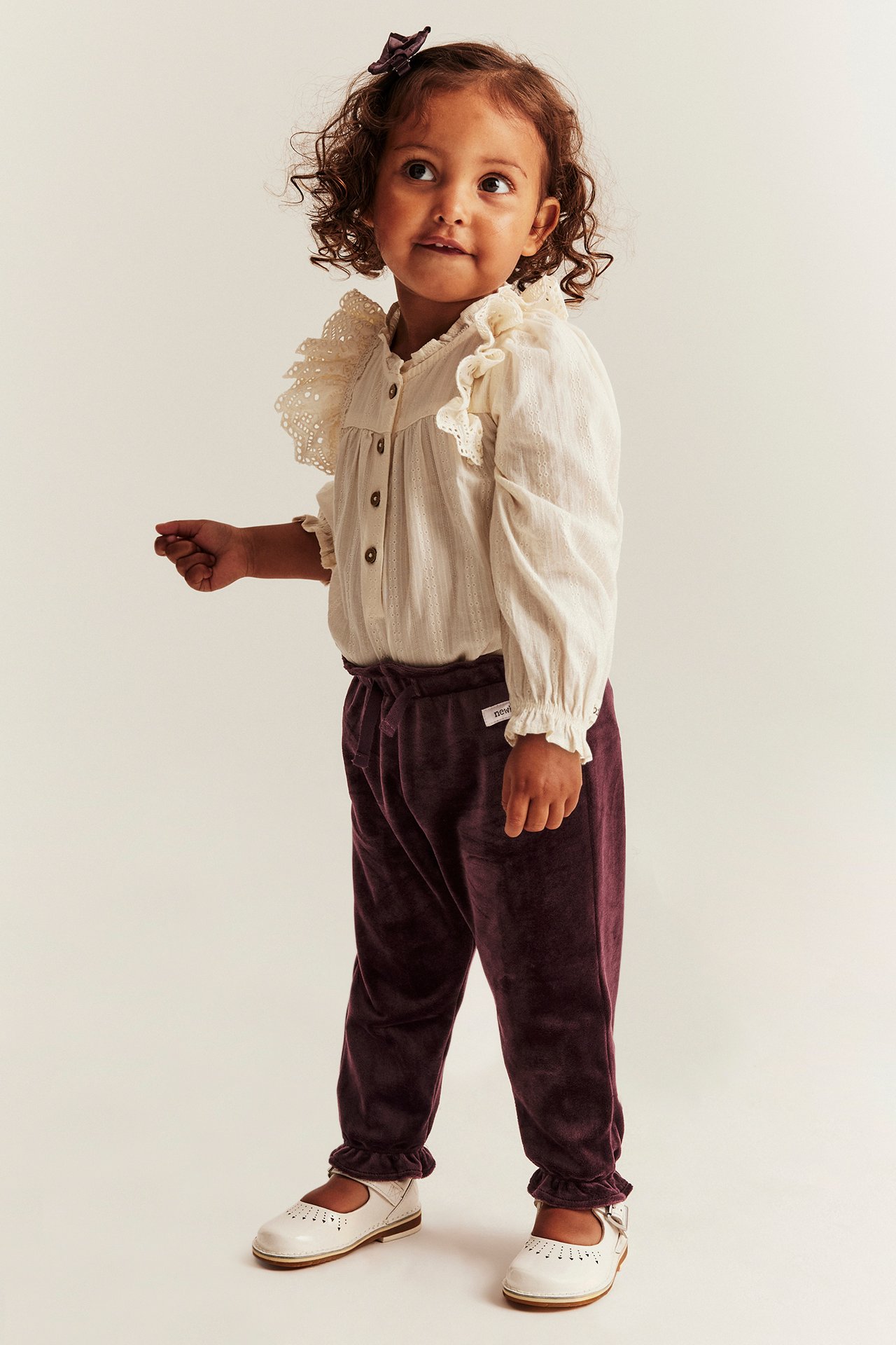 Front view: Kid in dark lilac velour jogging pants, off-white ruffled blouse, white shoes, dark hair clip.