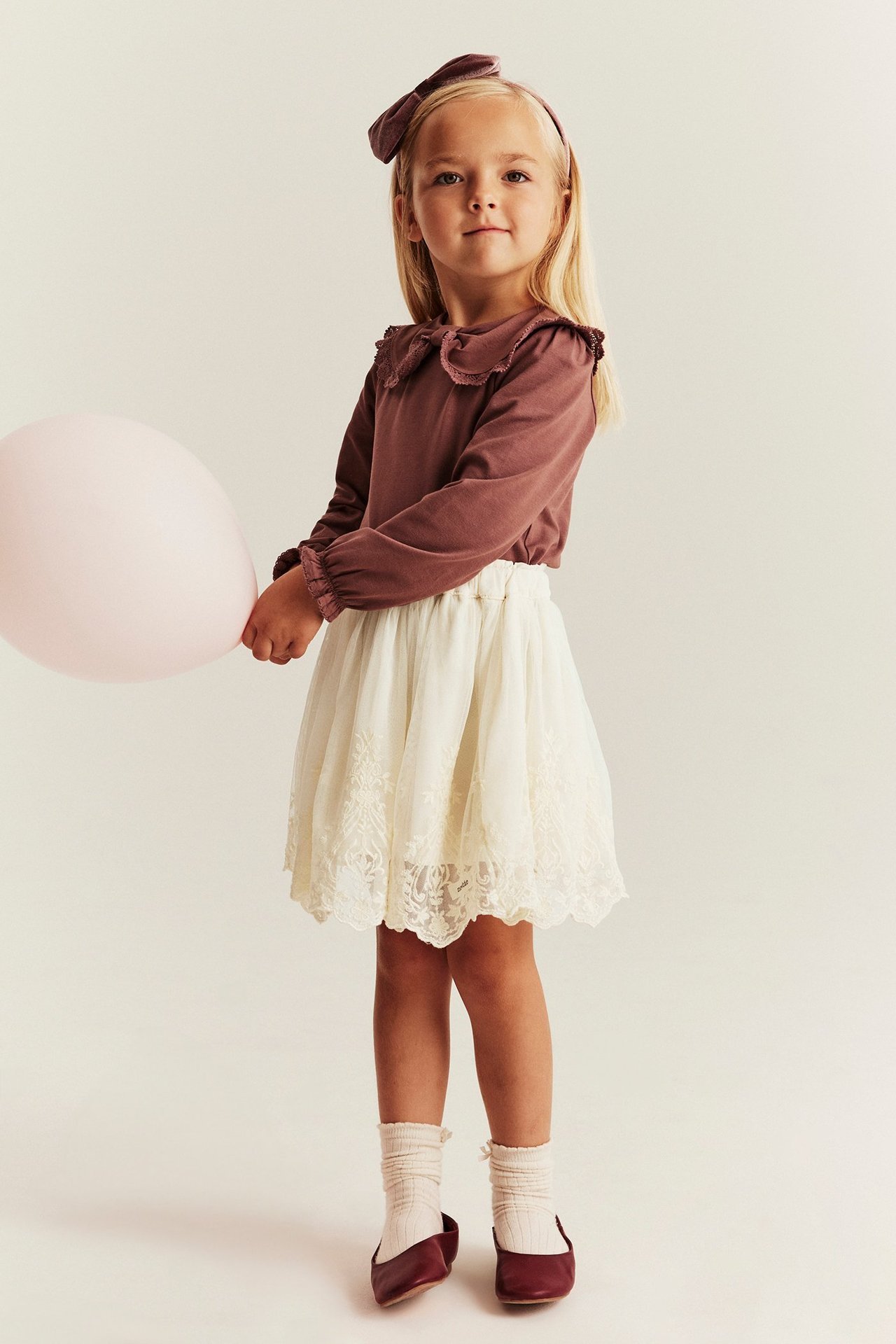 Kid in light beige embroidered mesh skirt, brown shirt, bow, cream socks, red flats, holding balloon.