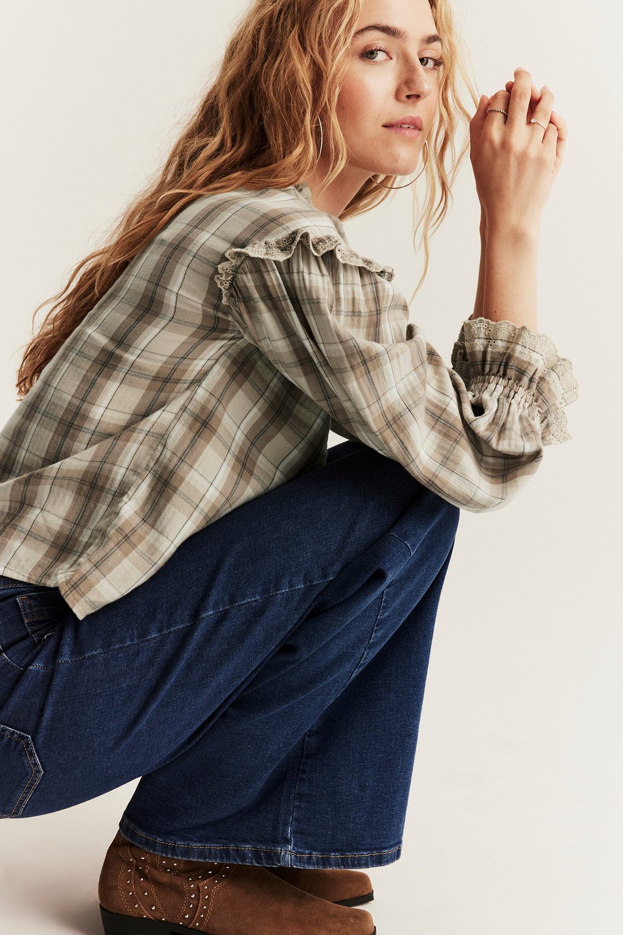 Angled front view of a woman in a beige plaid blouse with ruffles, dark blue jeans, and brown studded boots.