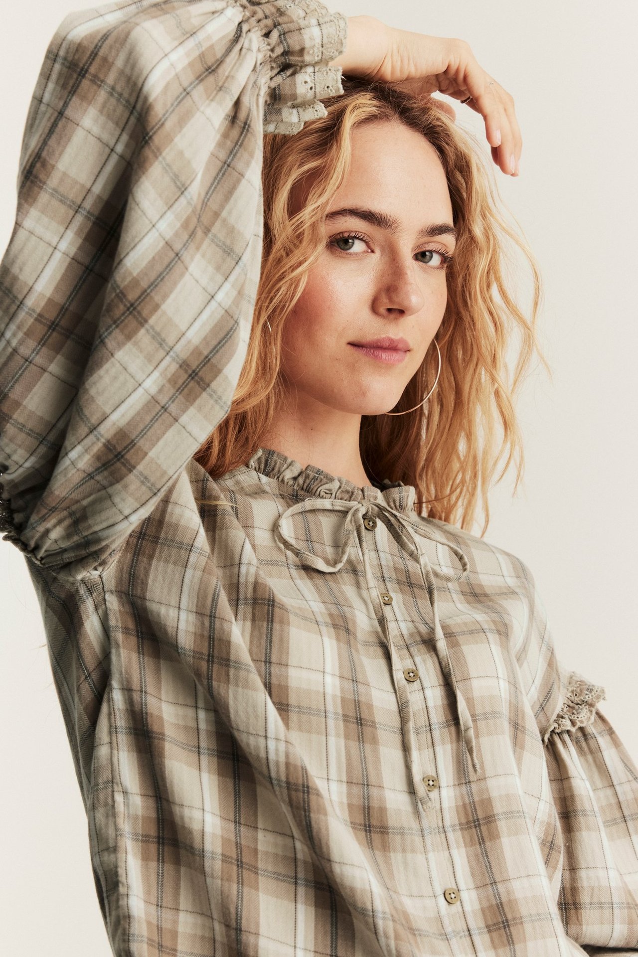 Nahaufnahme einer Frau in einer beigen karierten Baumwollbluse mit Rüschenausschnitt und bestickten Manschetten.