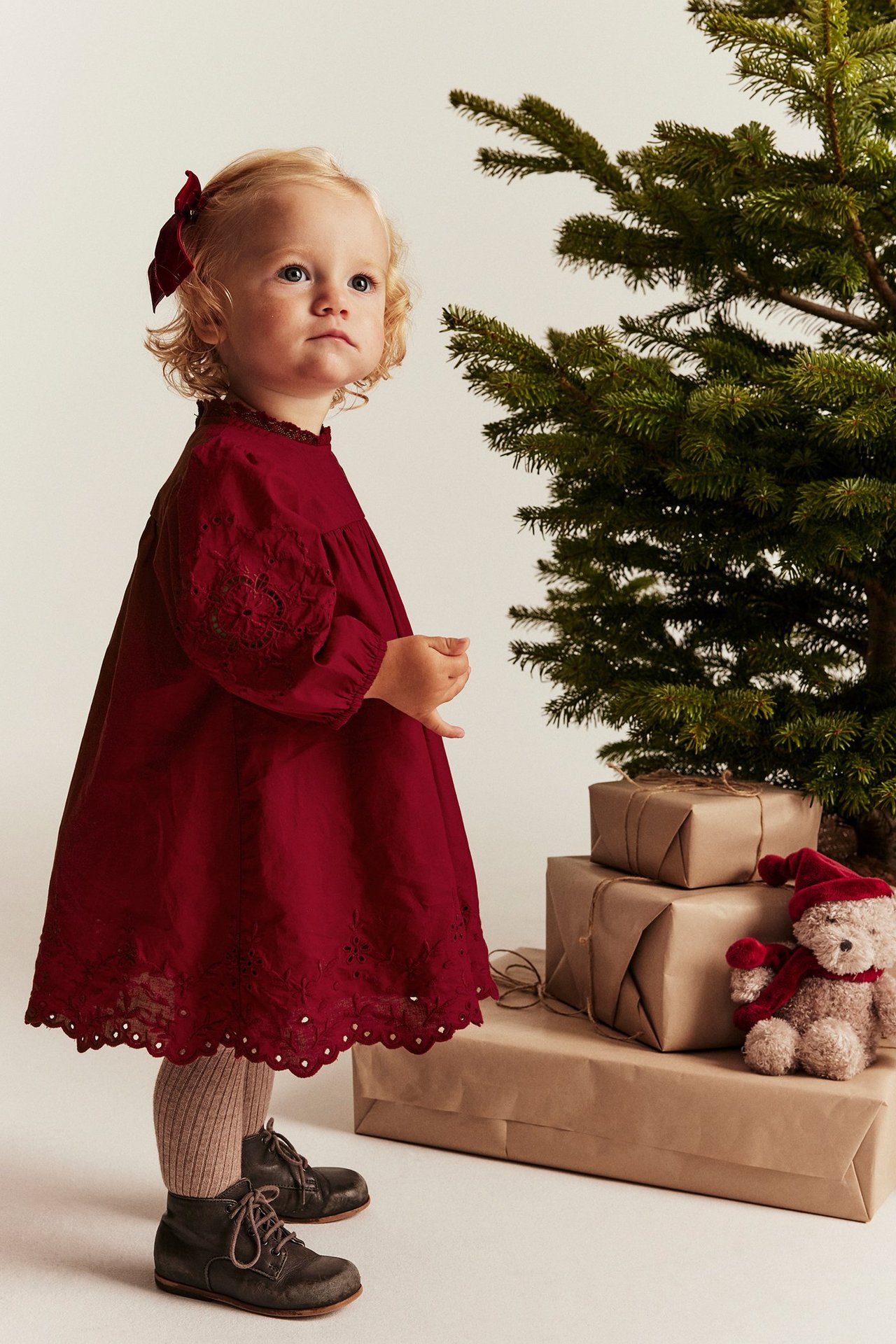 Baby girl in red embroidered cotton dress, brown tights, boots, and red bow by Christmas tree.