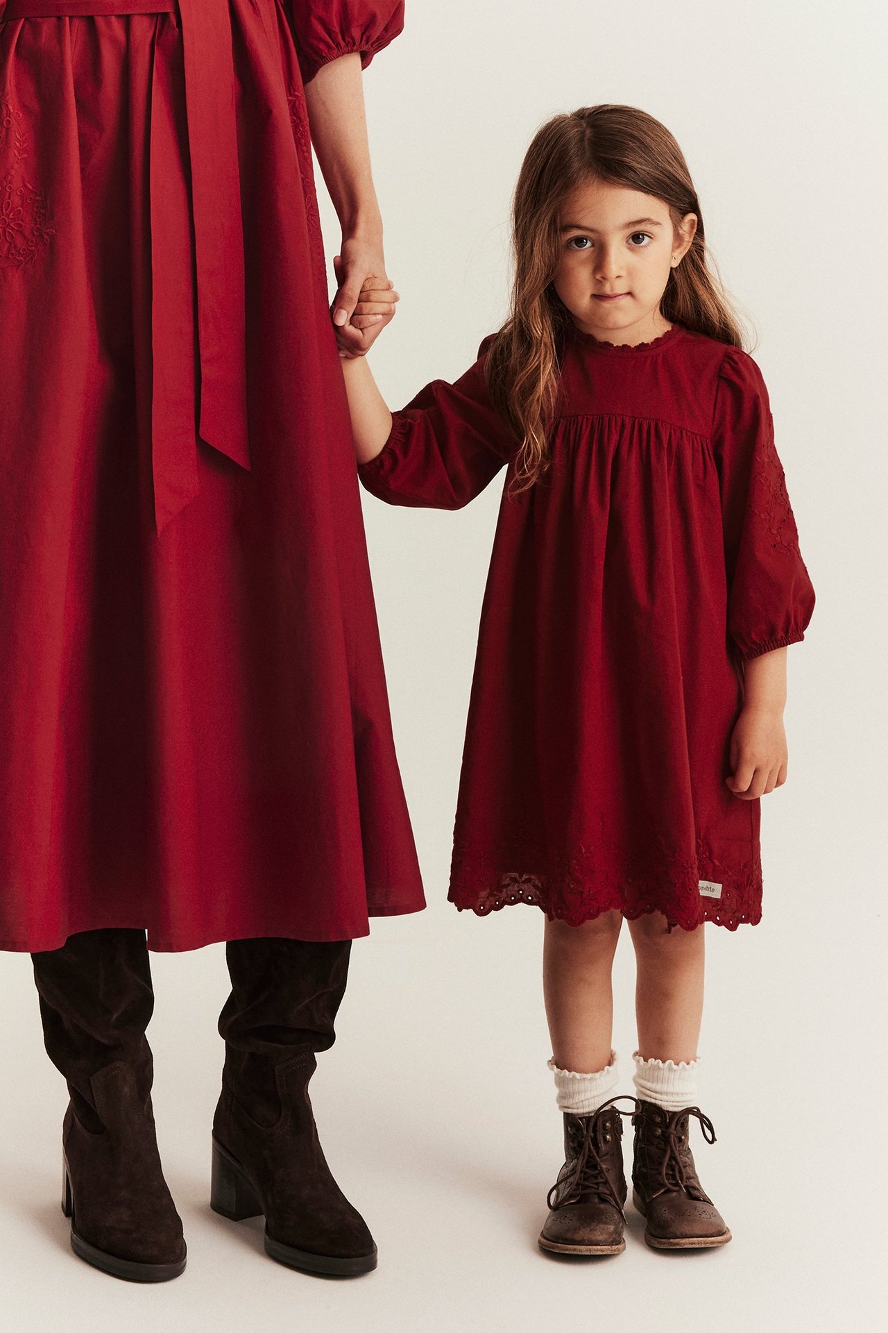 Front view of a kid in a red embroidered cotton dress with puff sleeves, holding a woman's hand.